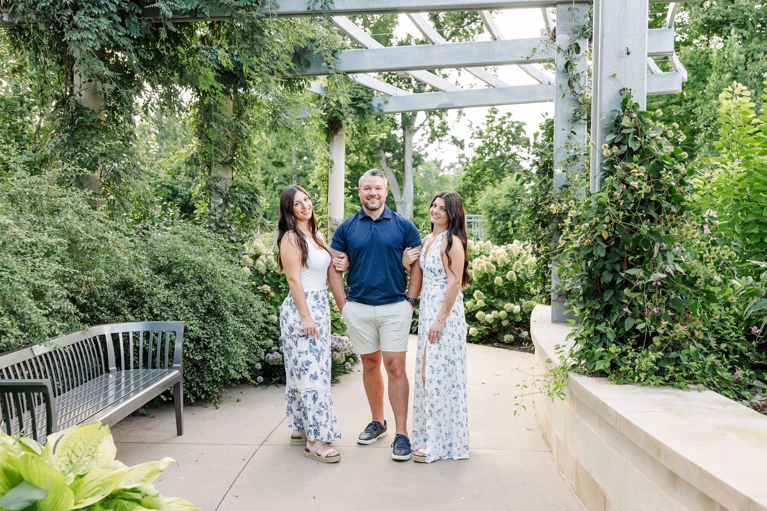 A man in navy polo and khaki shorts stands smiling between two women in matching blue floral maxi dresses in a garden.