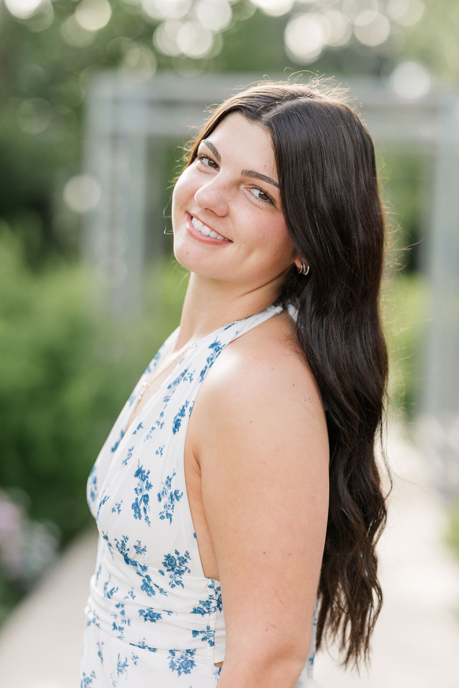 Close-up portrait of smiling brunette woman in blue floral dress in bright garden setting.