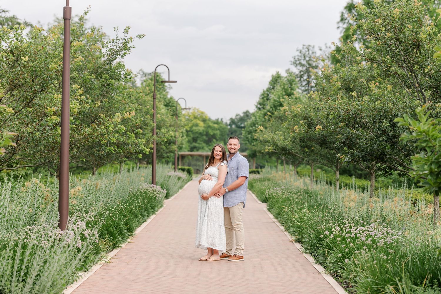 Pregnant couple poses on a brick garden path surrounded by lush greenery and wildflowers during a maternity photo session.
