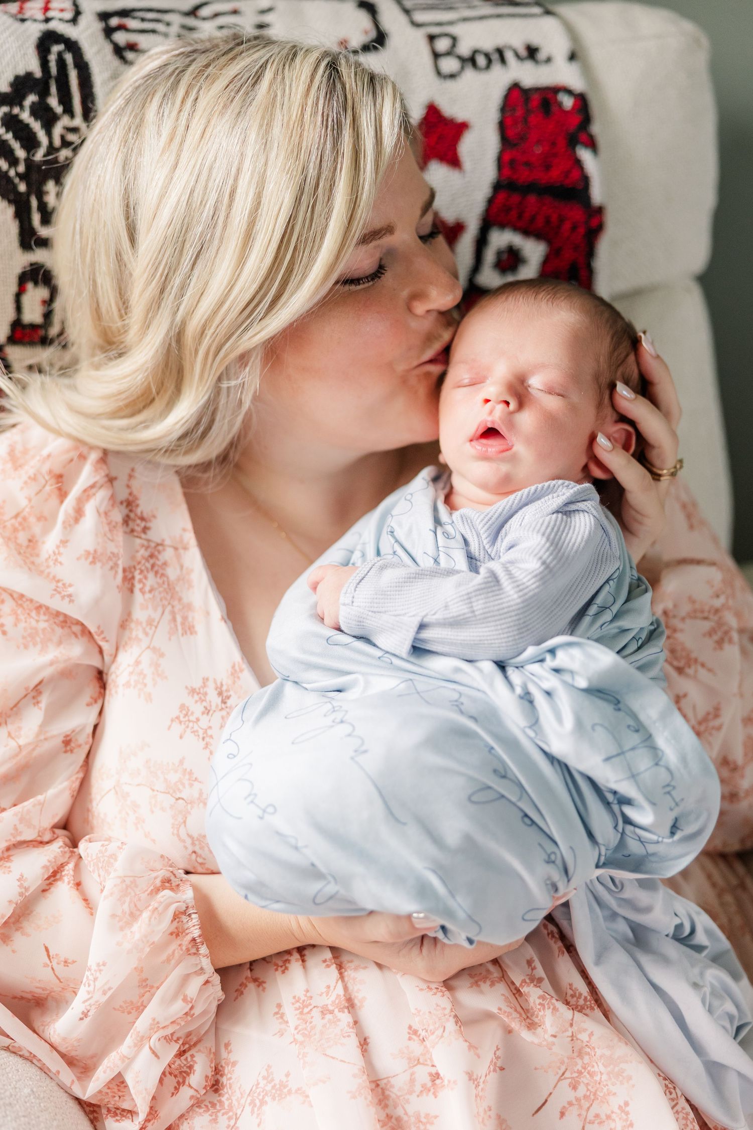 Blonde mother in pink floral dress kissing newborn baby dressed in light blue outfit.