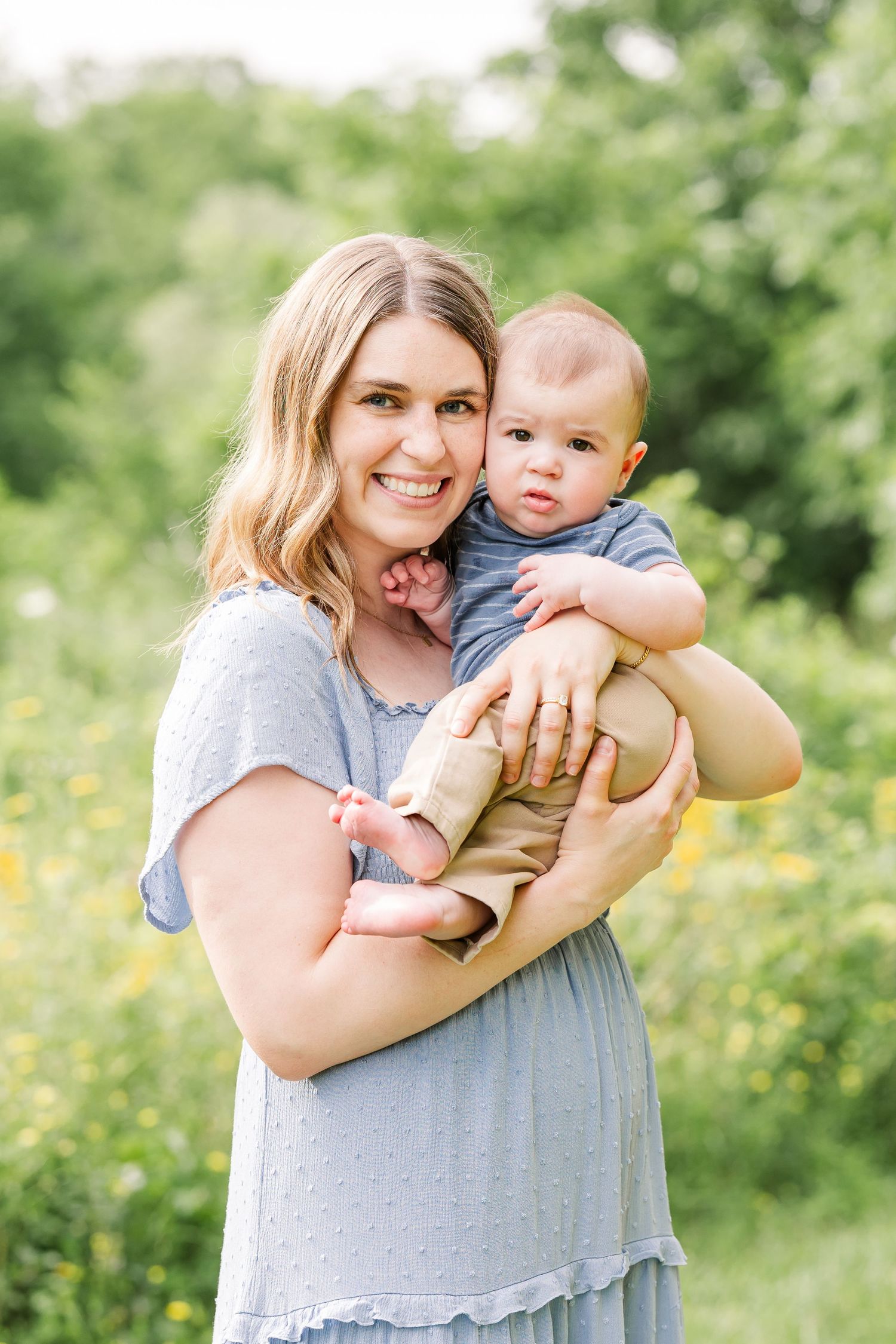 Smiling mother in blue dress holds her infant baby outdoors in a lush green field with yellow wildflowers.