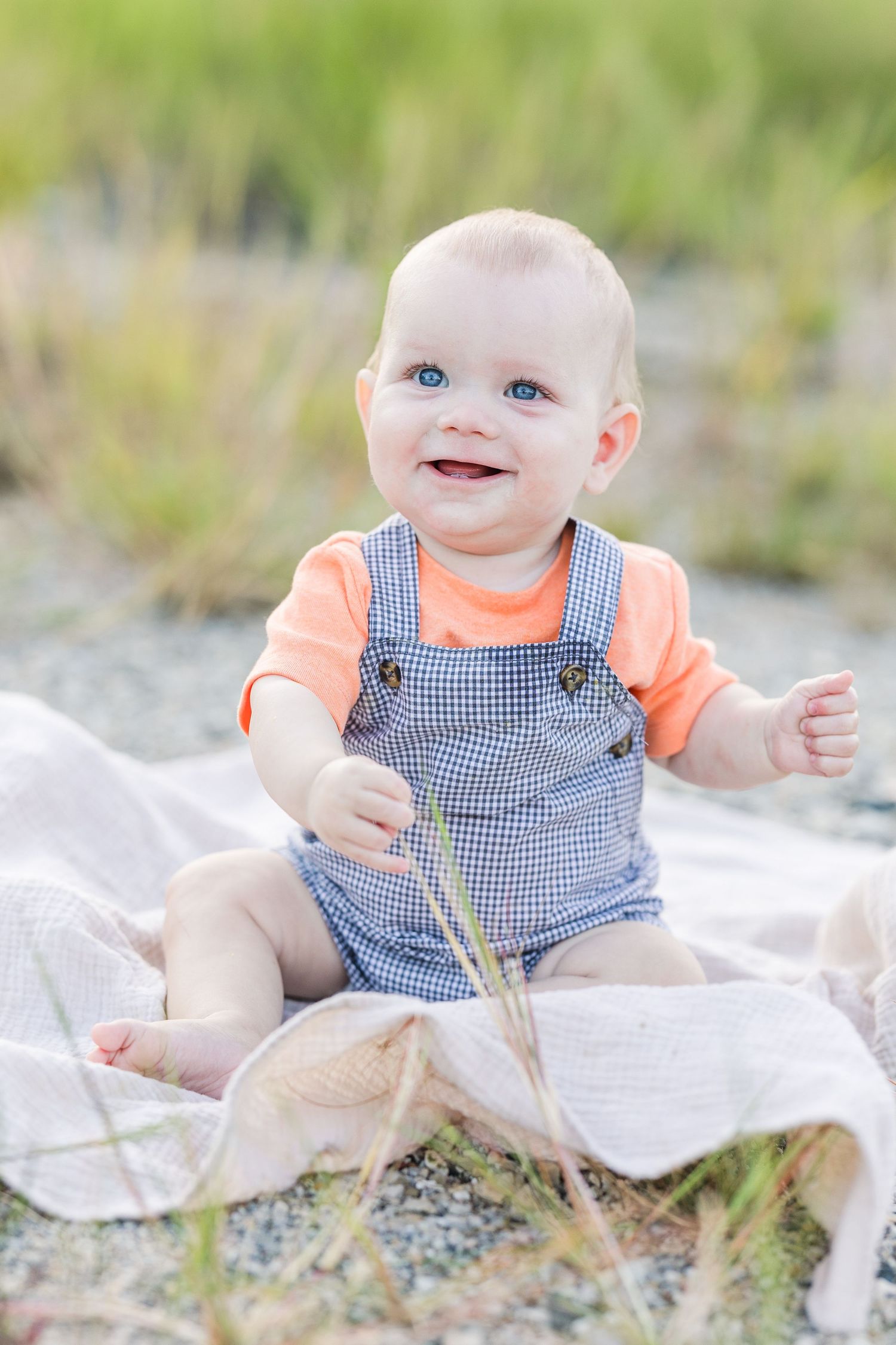 Baby wearing orange shirt and blue checkered overalls sits on white blanket outdoors in grassy area.