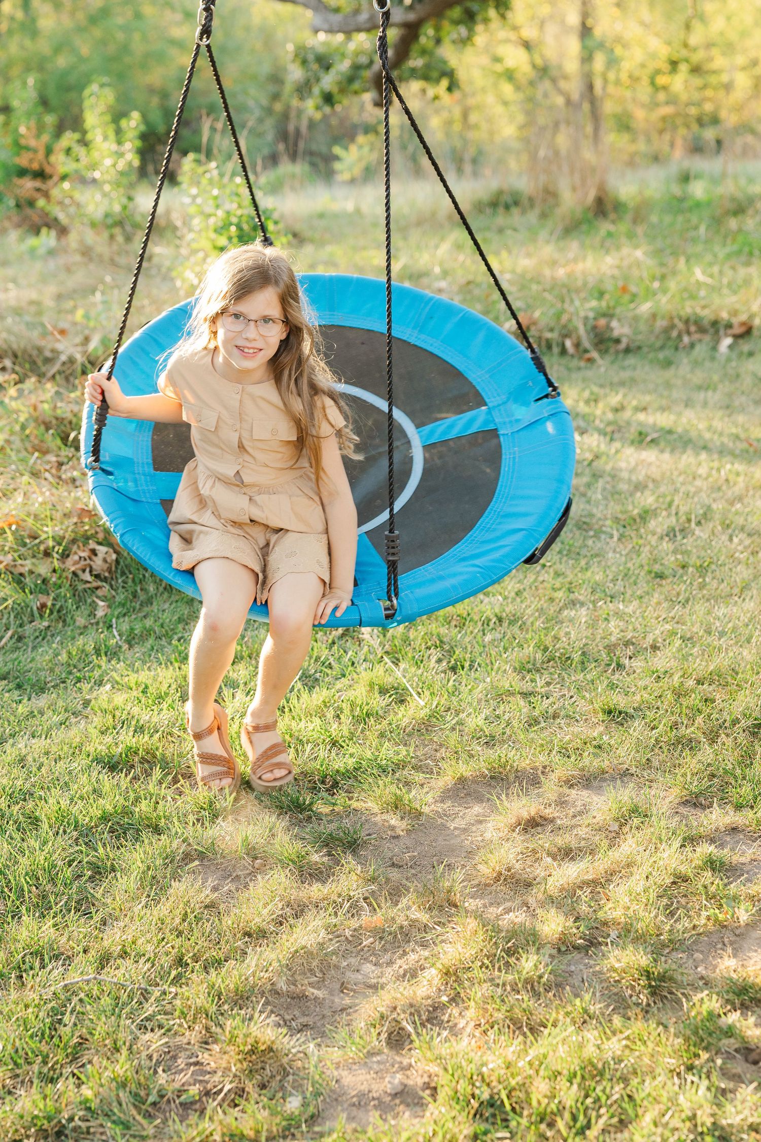 A blue disc tree swing sways gently in a sunny backyard setting with green grass and natural lighting.