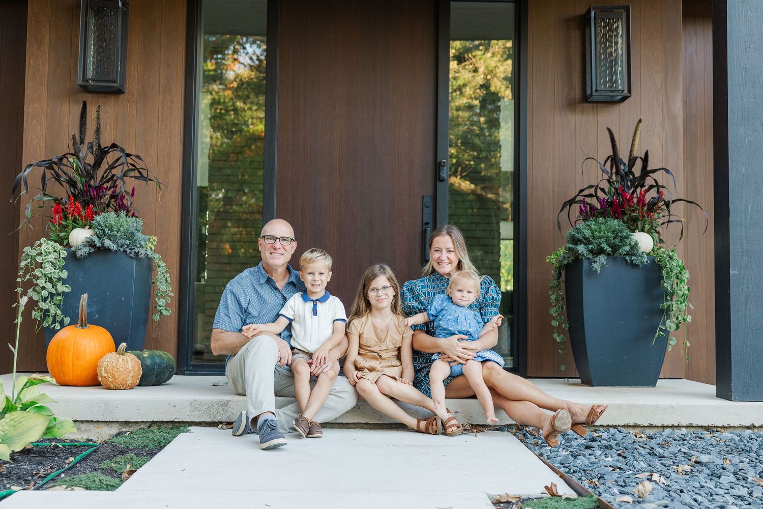 Family sitting on front porch steps decorated with pumpkins and fall decor during autumn season.