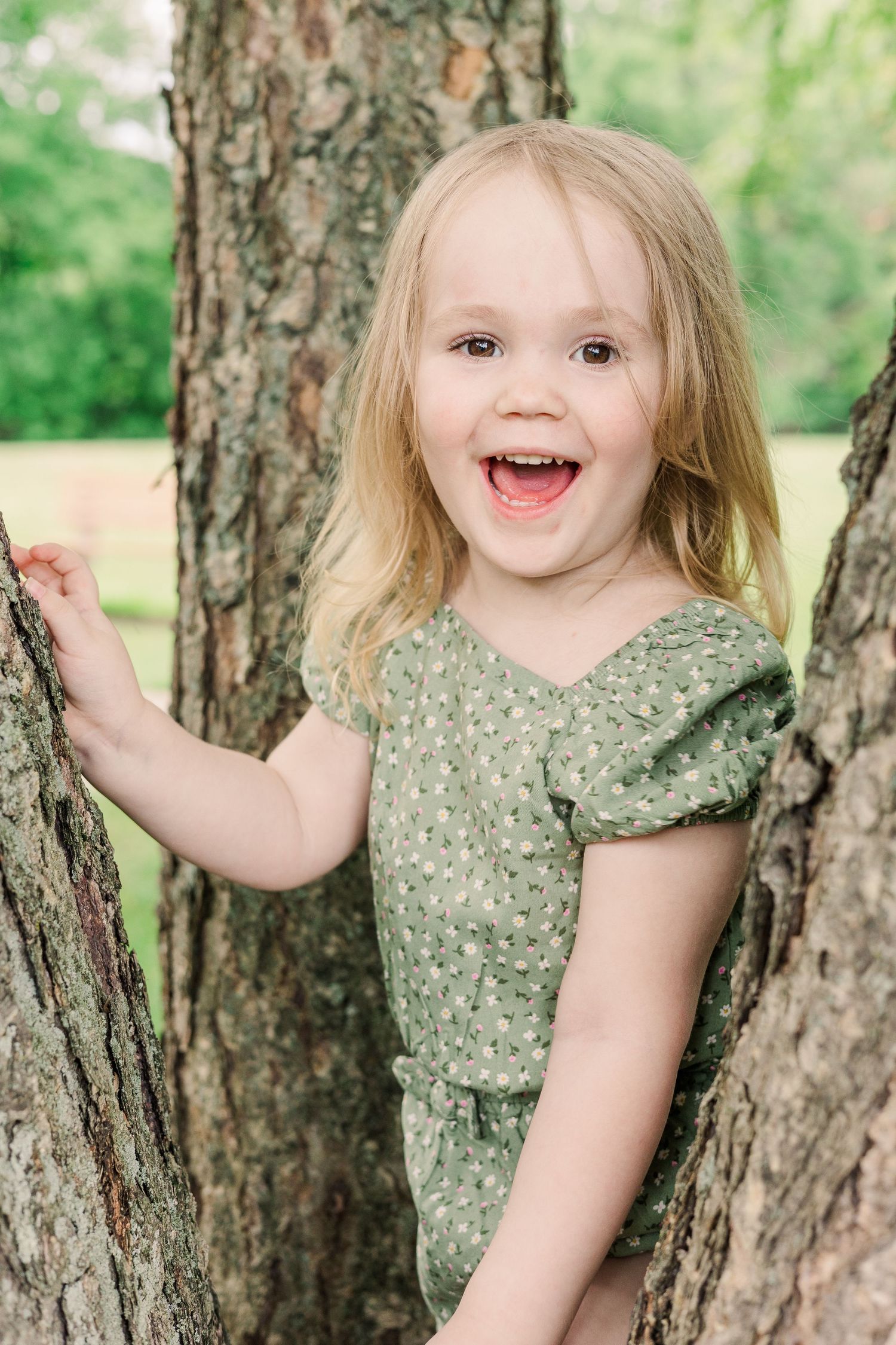 A joyful child in a green dress laughs while playing between two trees in a park.