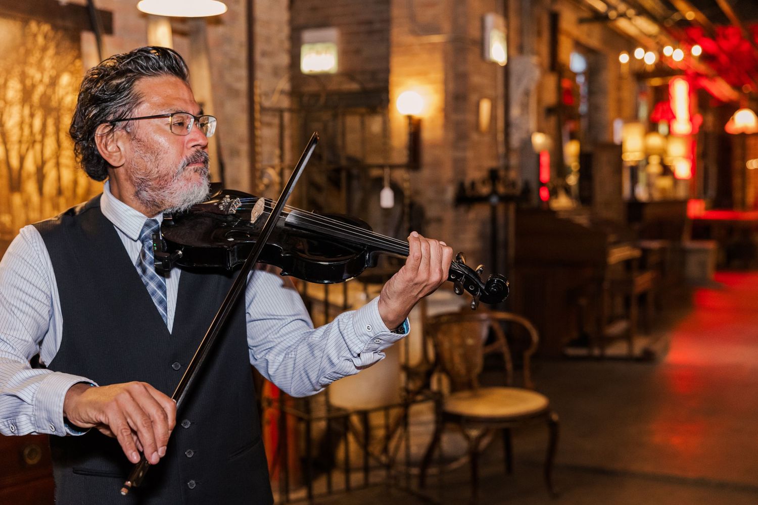 A violinist in a vest and bowtie performs in a dimly lit venue with exposed brick walls.