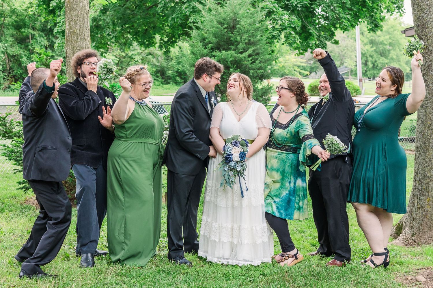 Wedding party in green dresses poses together outdoors while celebrating enthusiastically.