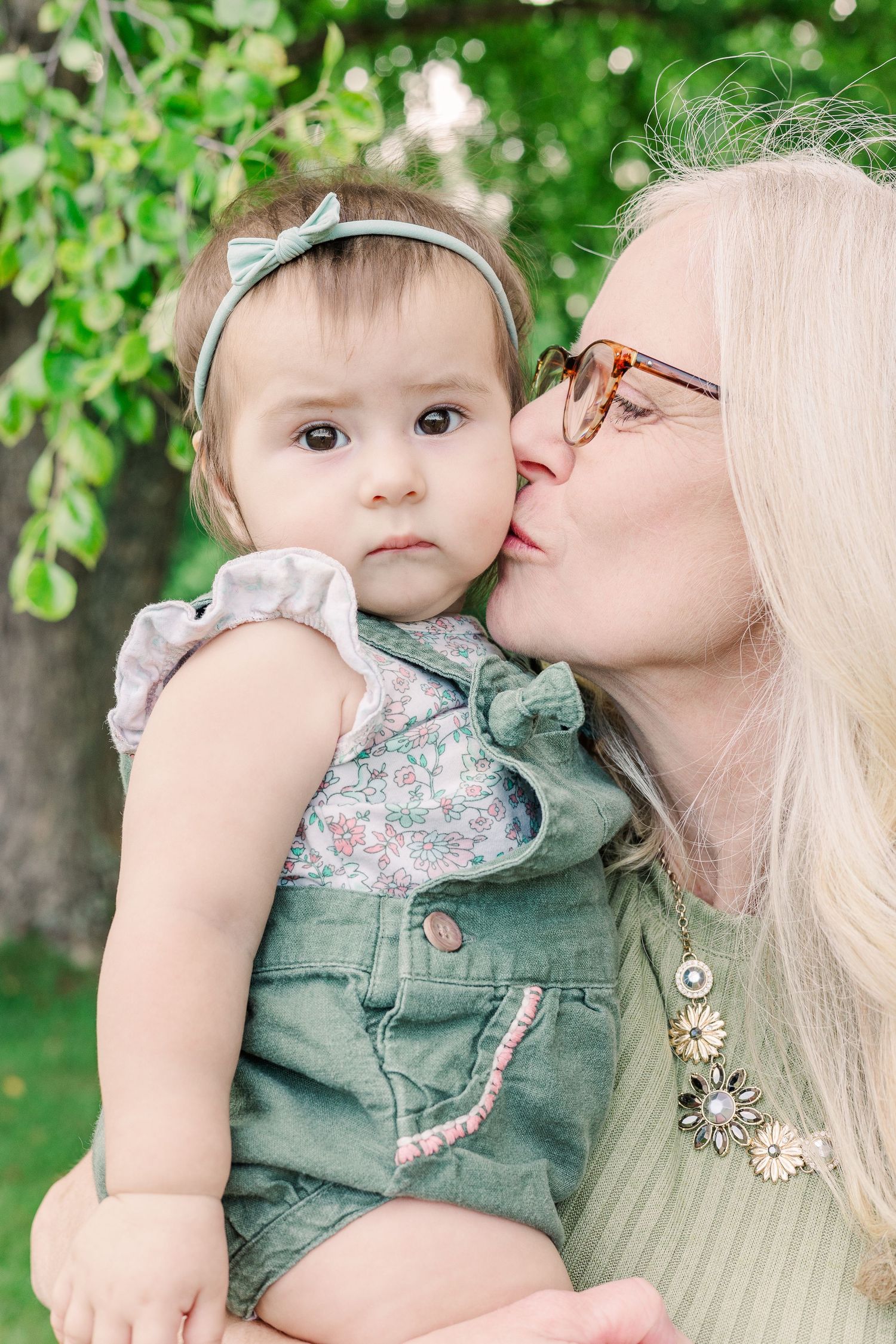 Close-up portrait of parent giving baby in green overalls a gentle kiss outdoors.