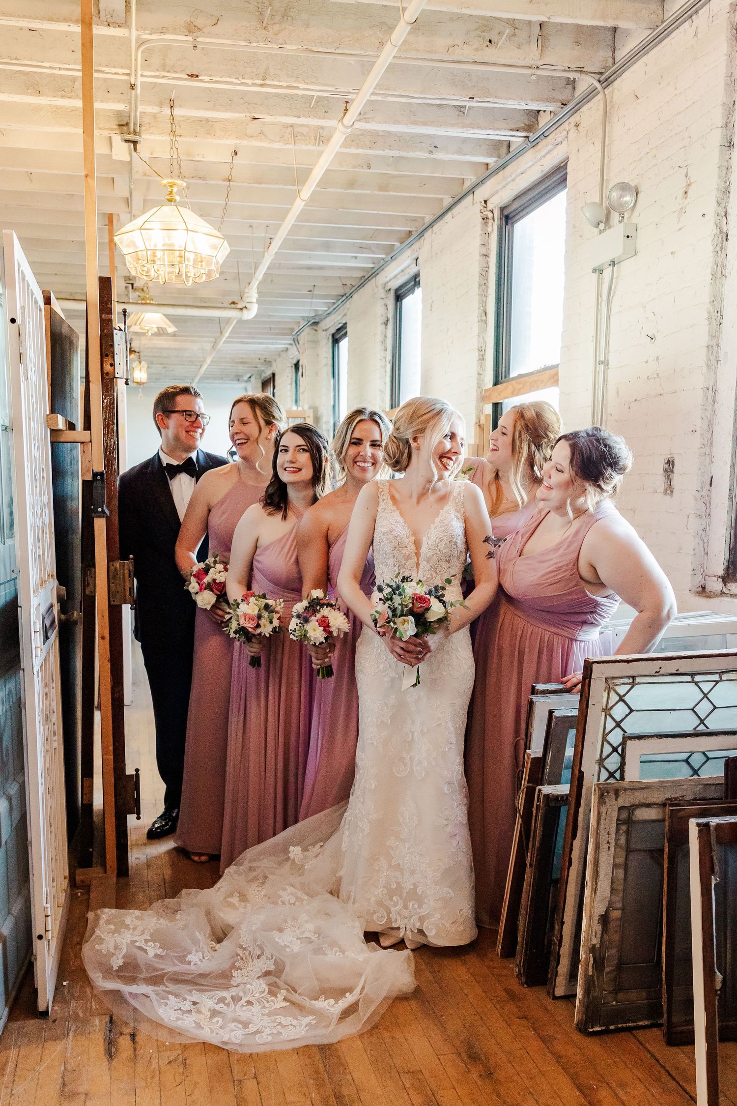 Bridal party gathers in industrial chic hallway with wooden floors and crystal chandelier overhead.