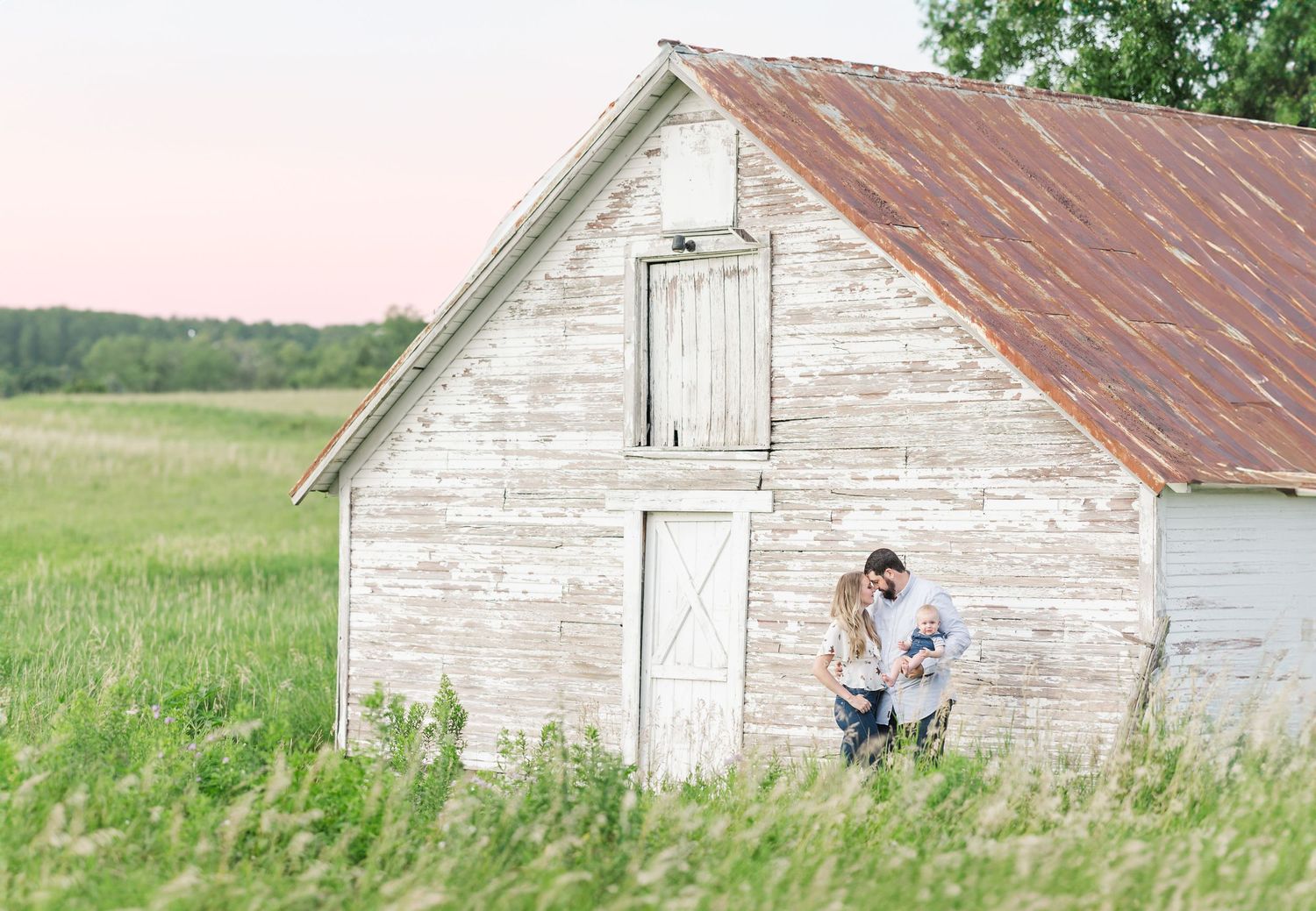 A tender moment shared beside a weathered white barn in a rolling field at dusk.