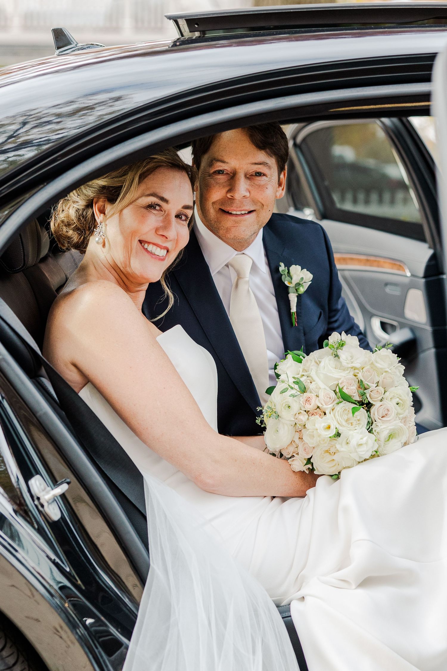 Wedding couple sitting in black car with white bouquet and formal attire.