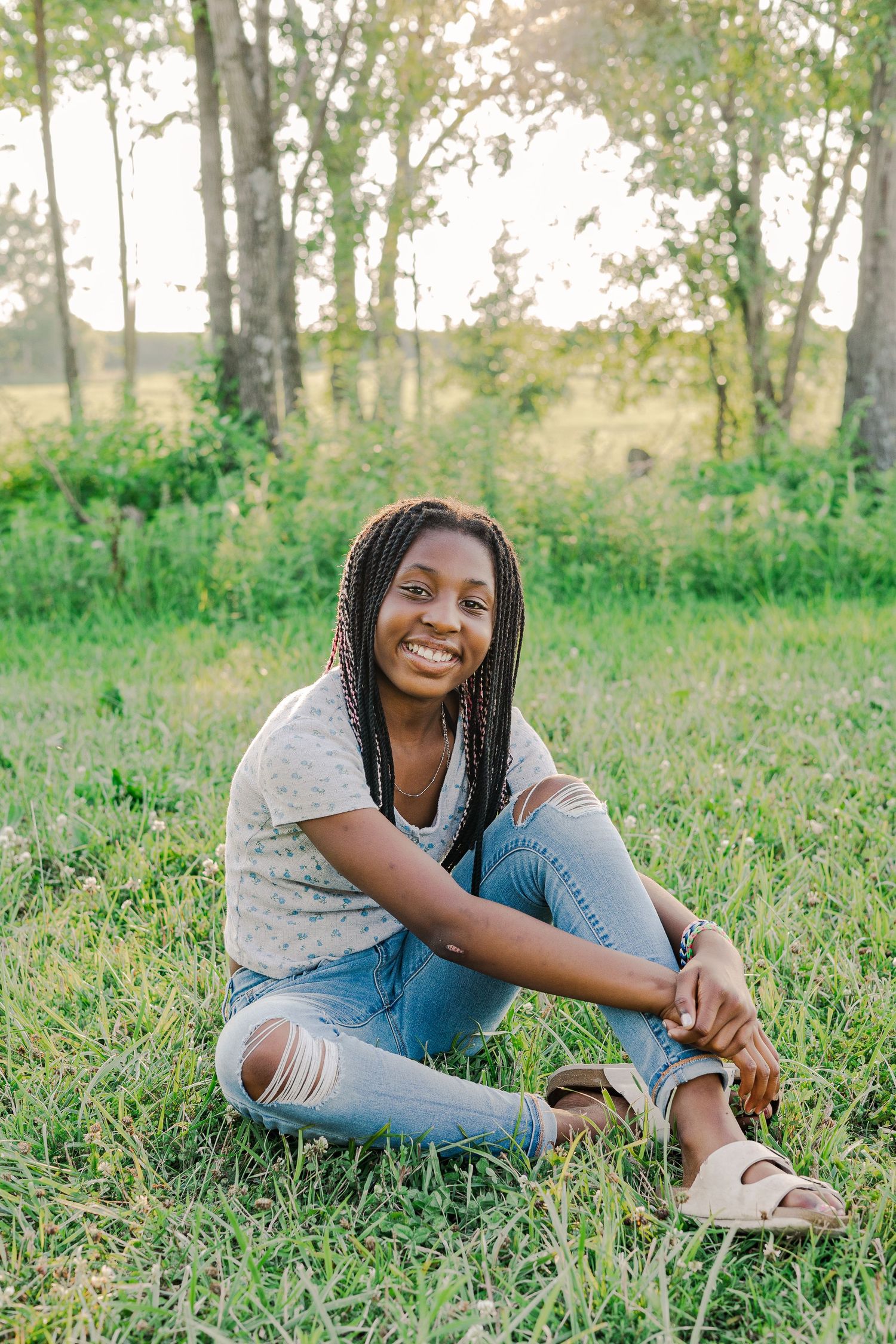 Someone sitting in ripped jeans on grass with a bright smile against a nature backdrop.