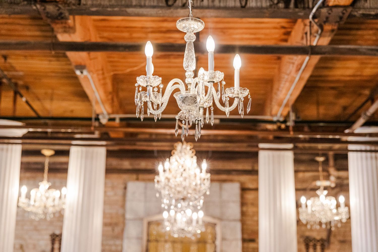 Elegant crystal chandeliers hanging from a wooden beam ceiling in a rustic venue.