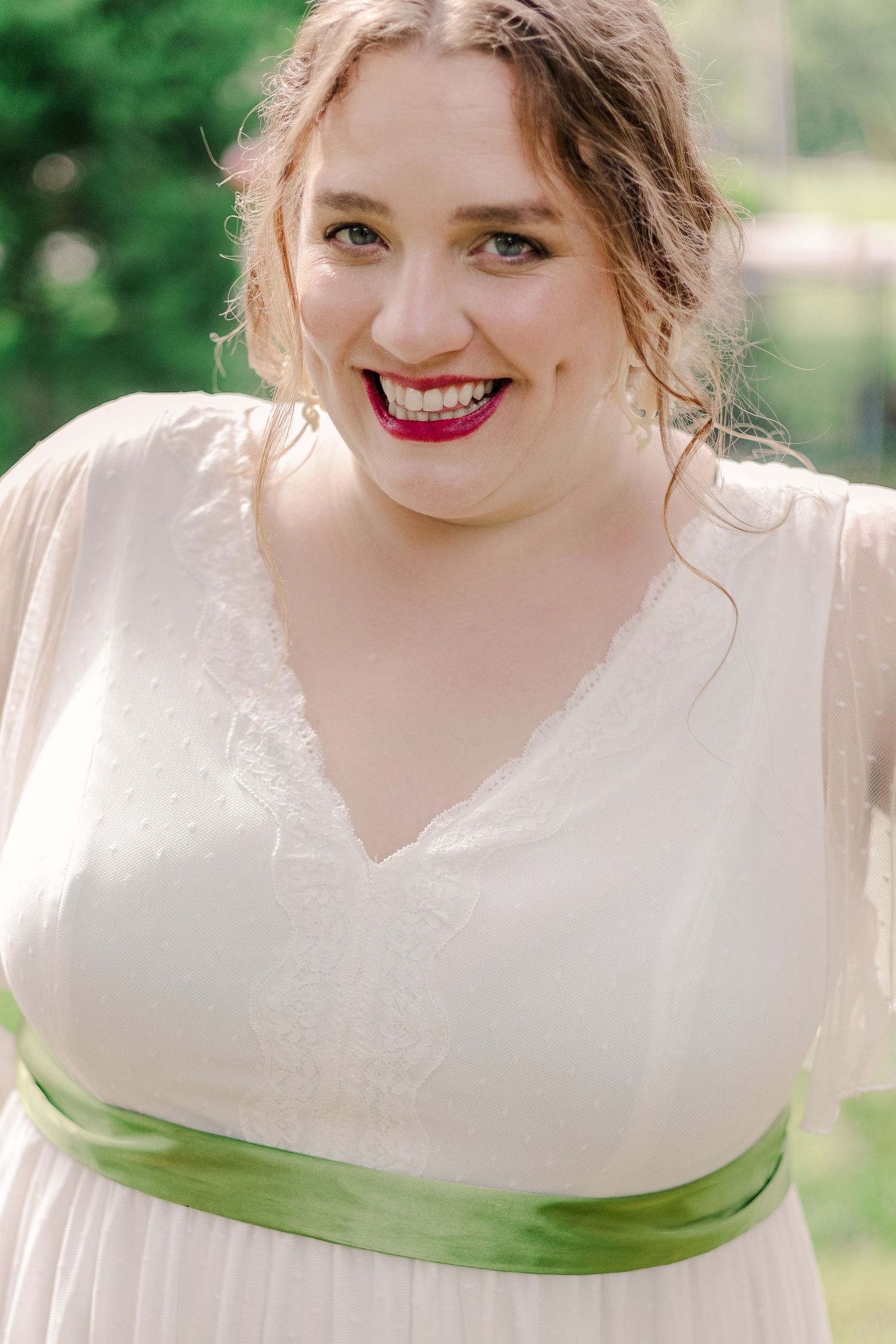 Bride in white dress with green sash and bold lipstick smiles outdoors.
