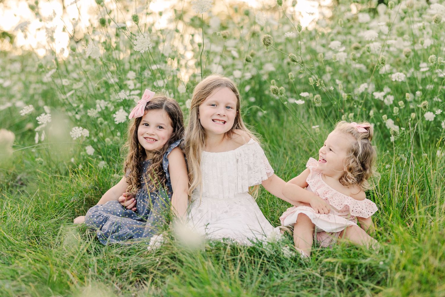 Three people sitting together and laughing in a lush meadow filled with white wildflowers.