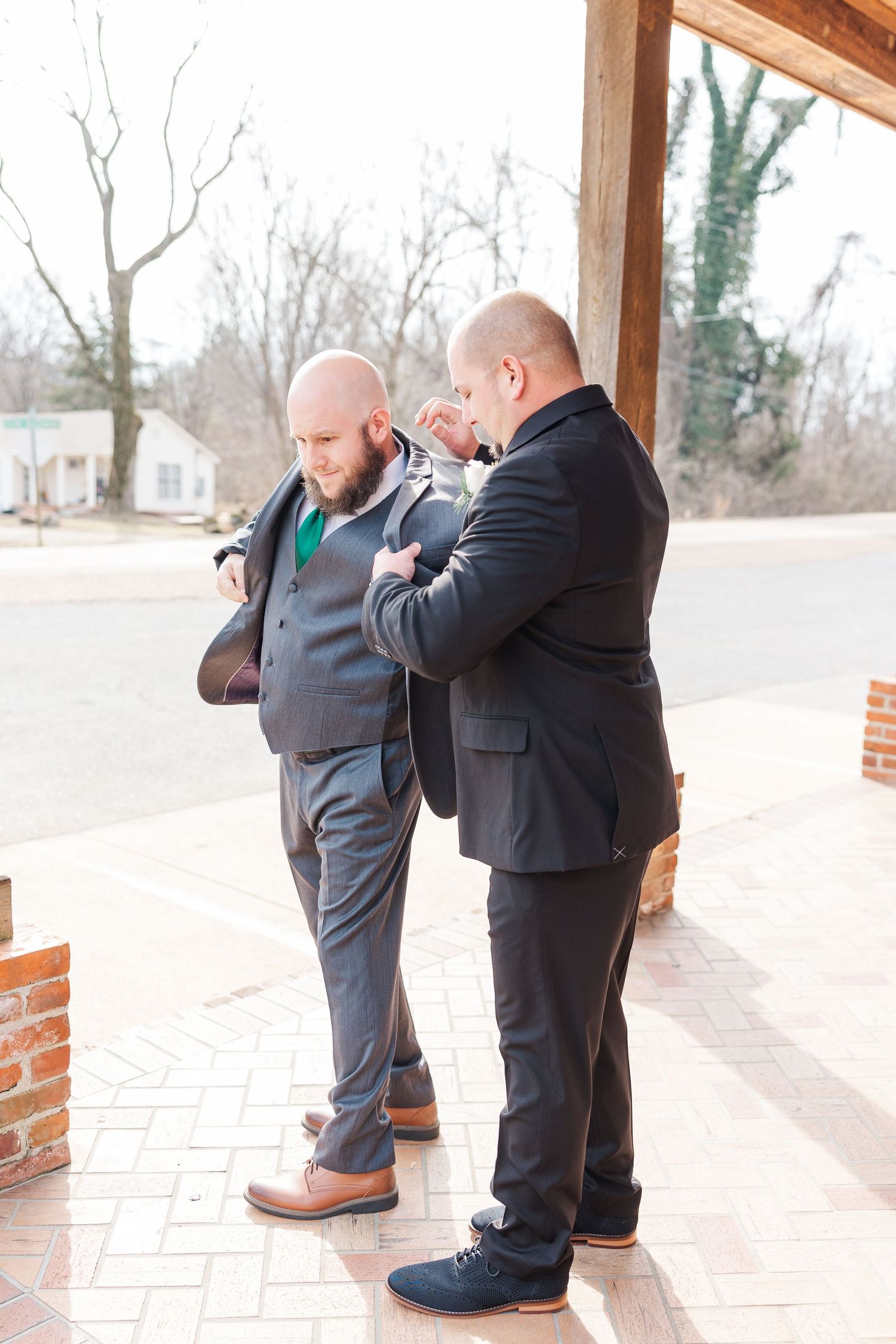 Two people in formal suits adjusting ties on covered porch.
