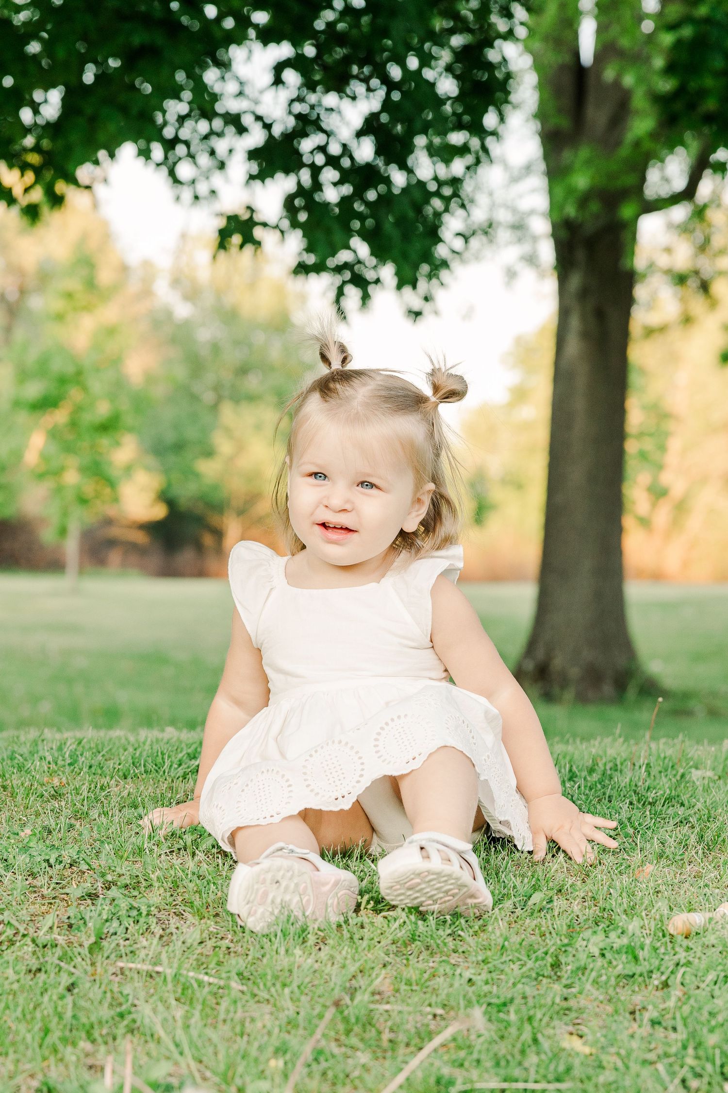 Young child in a white dress sitting on grass under a tree in summer.