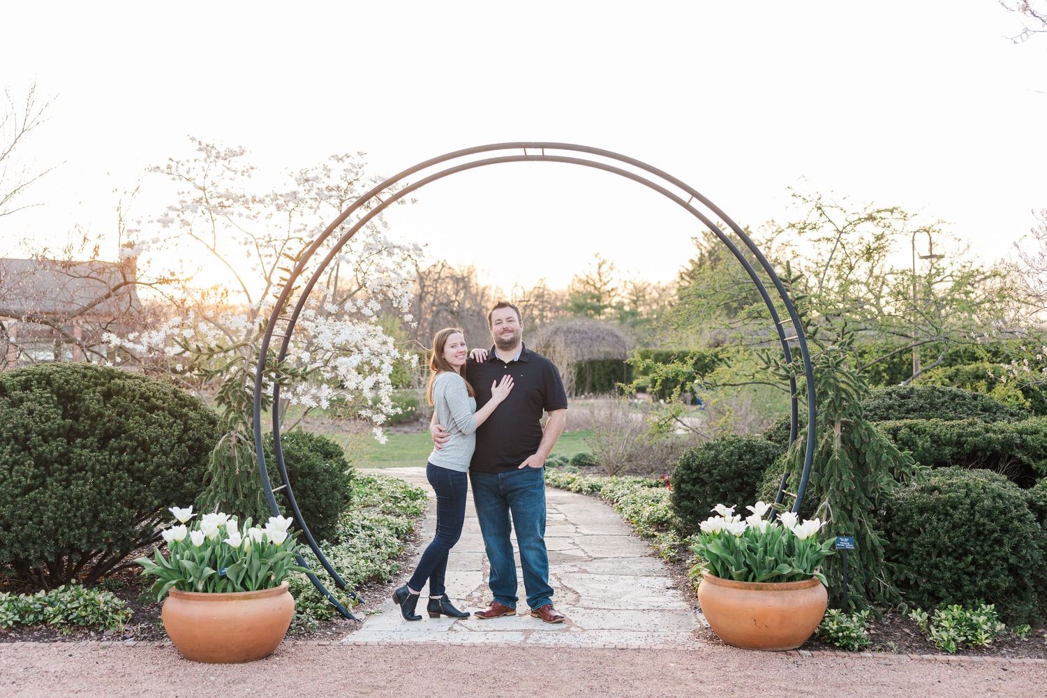 Couple standing together under decorative garden arch with potted plants and white flowers.