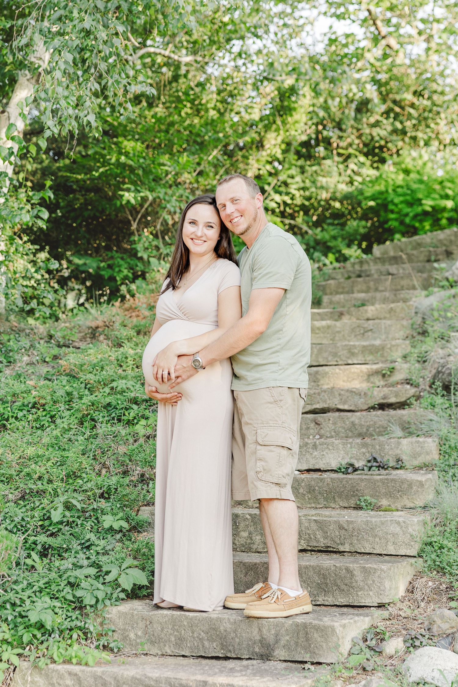 A couple stands on stone steps in a lush garden setting, with the woman wearing a flowing beige maternity dress.