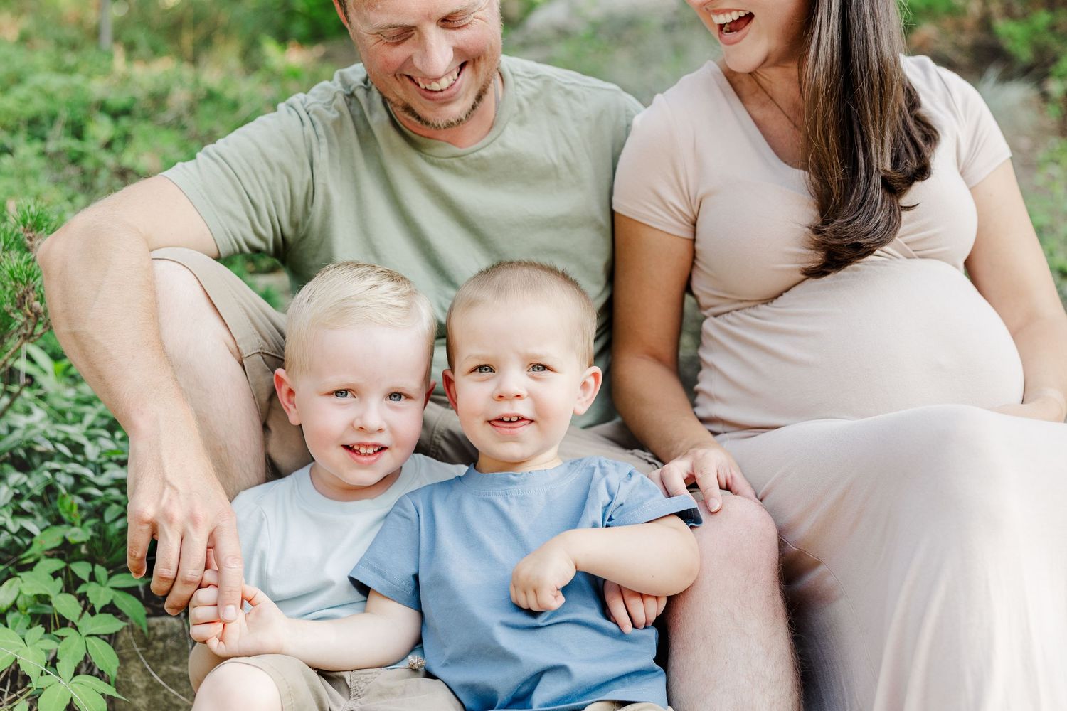 A joyful family moment outdoors with two young twins in matching outfits sitting in front of their parents in nature.