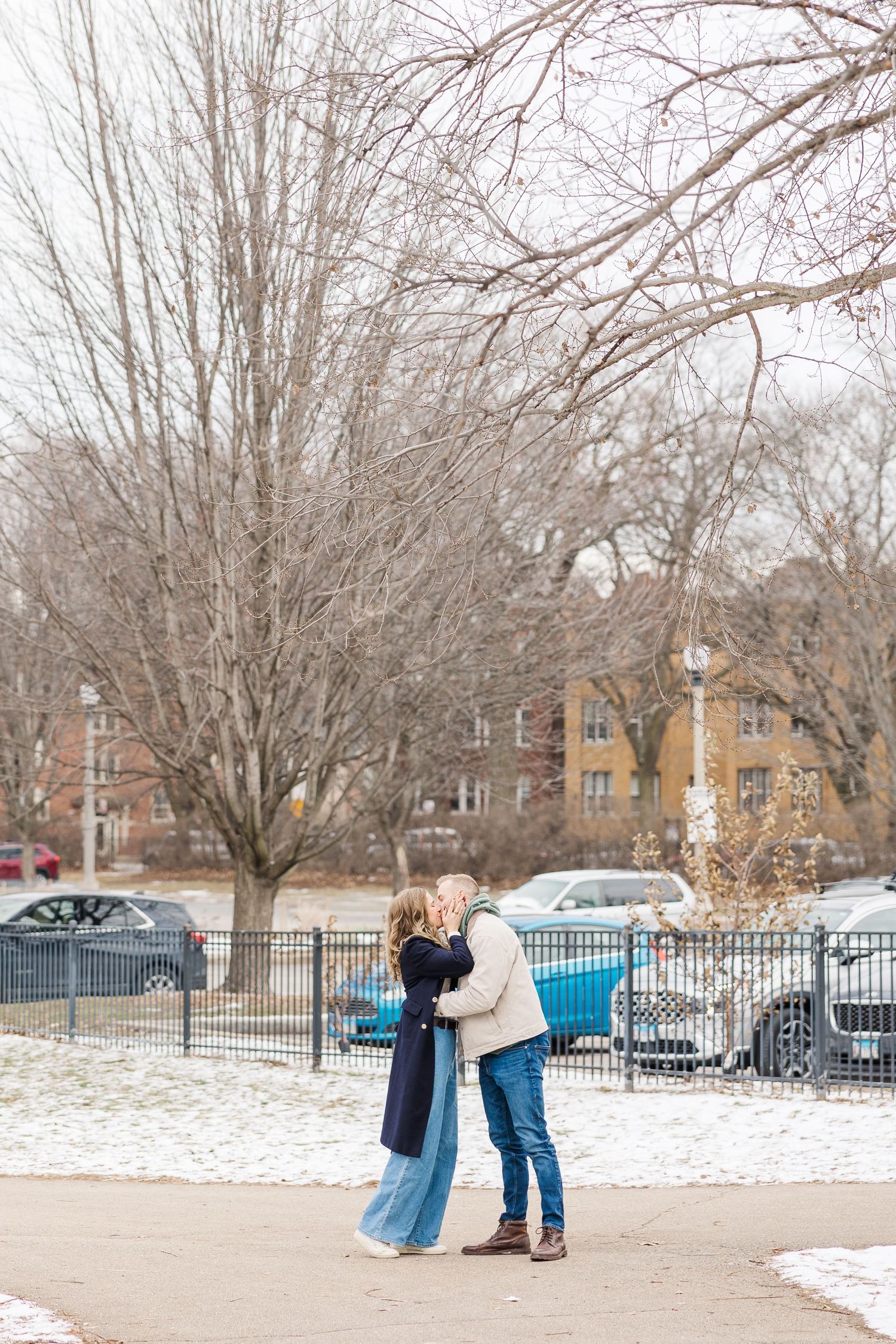 Man proposes to woman in snow-dusted park with bare trees and parked cars visible behind an iron fence in winter. Winnemac Park, Chicago