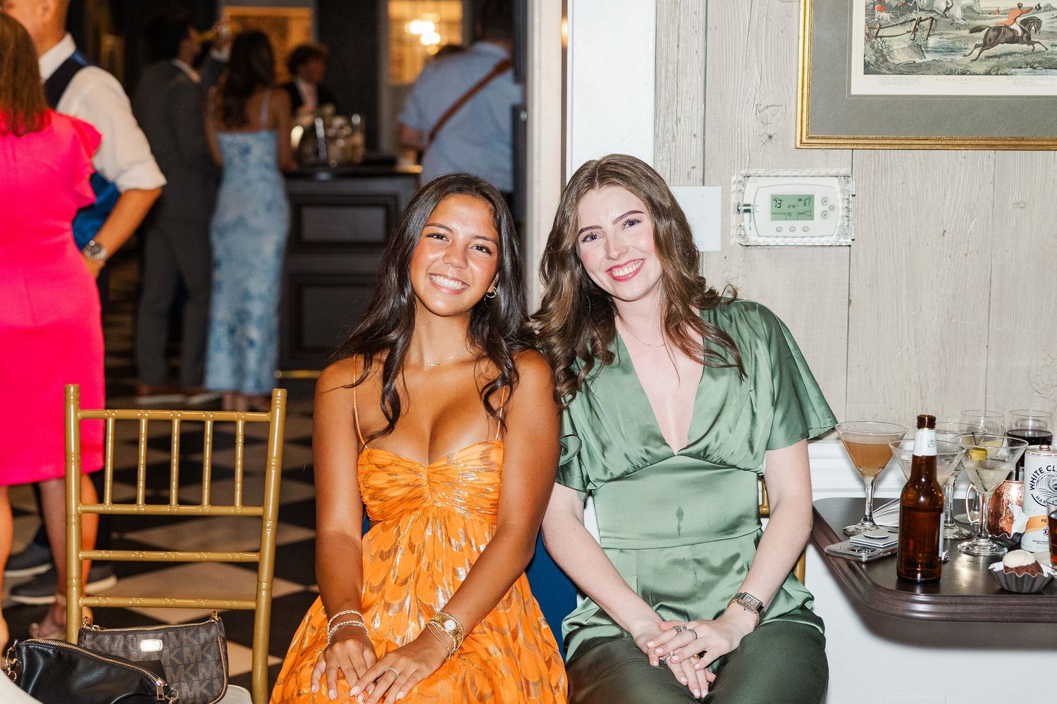 Two guests in orange and green dresses sharing a moment at a wedding reception table.