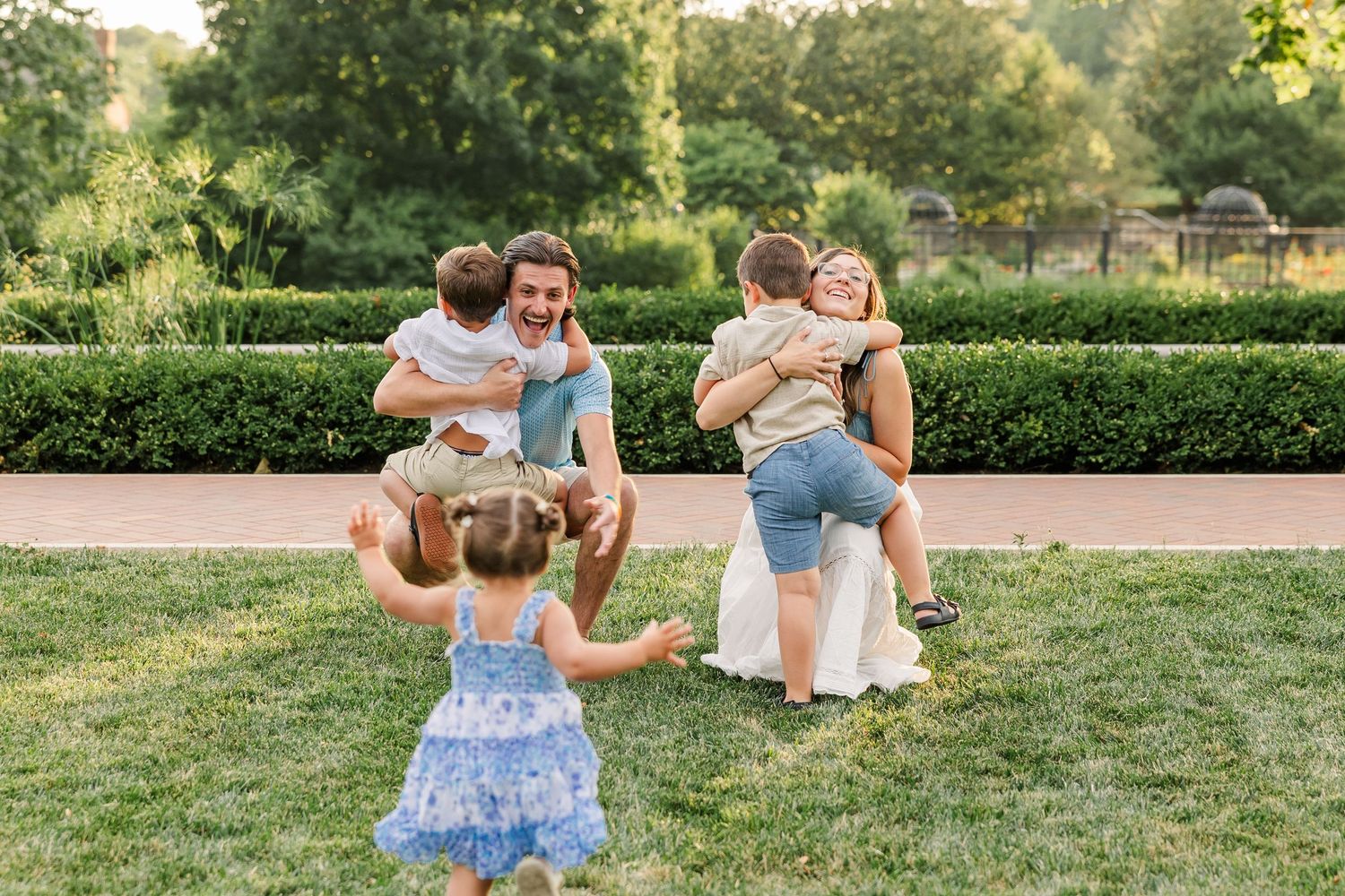 Family joyfully reuniting outdoors, parents hugging children on lush green lawn with manicured garden hedges in warm sunlight.