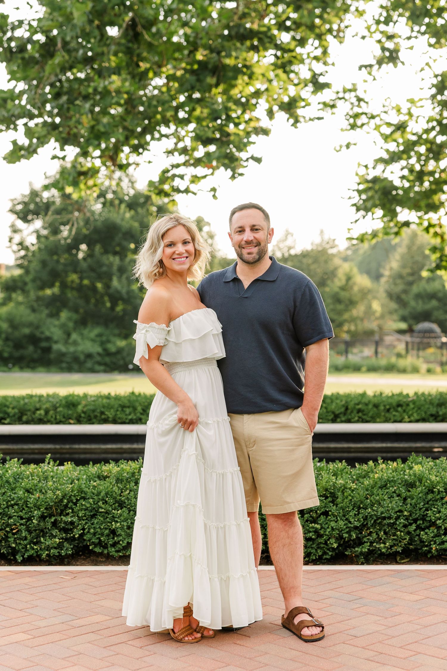 Smiling couple poses together on brick path in white dress and navy polo for outdoor portrait session.