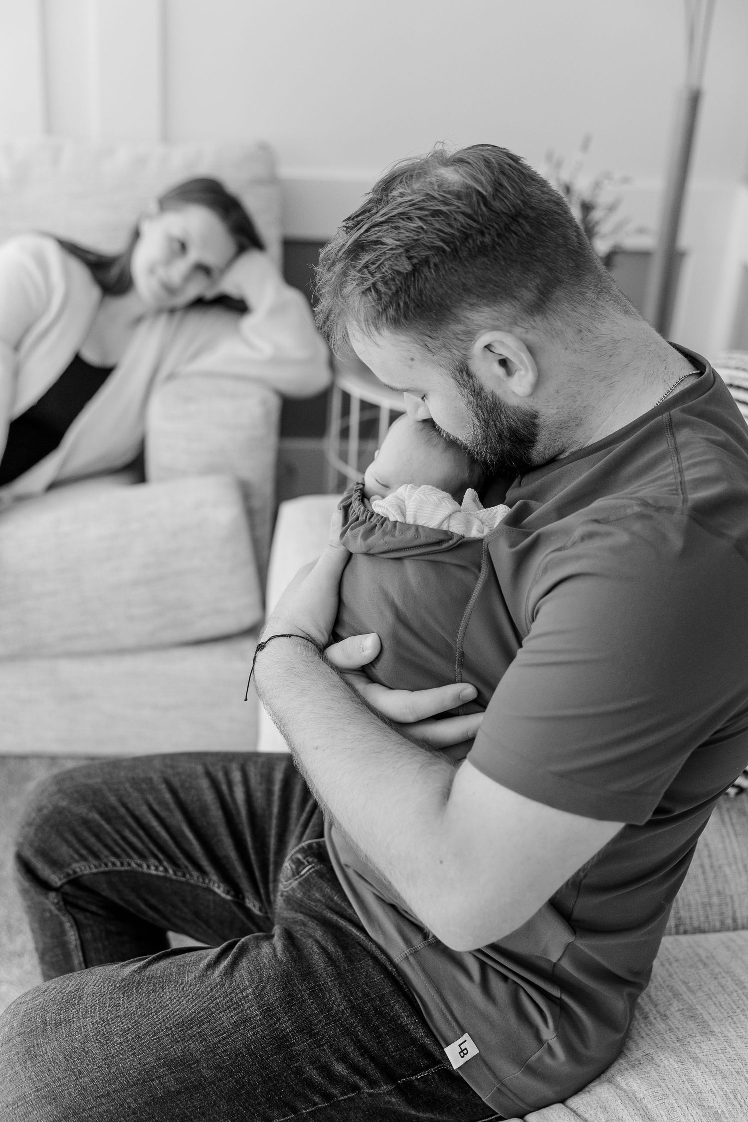 Black and white photo of bearded father tenderly kissing newborn baby while mother watches lovingly from sofa.
