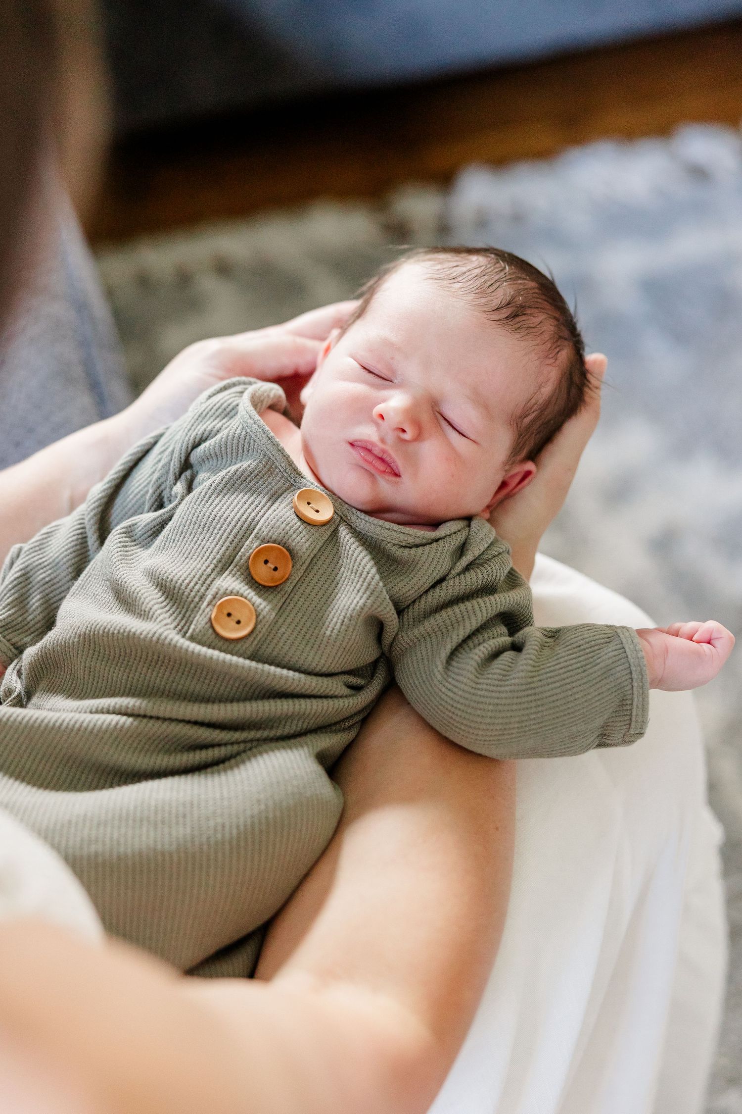 Newborn baby in sage green ribbed romper with brown buttons, peacefully resting in parent's arms.