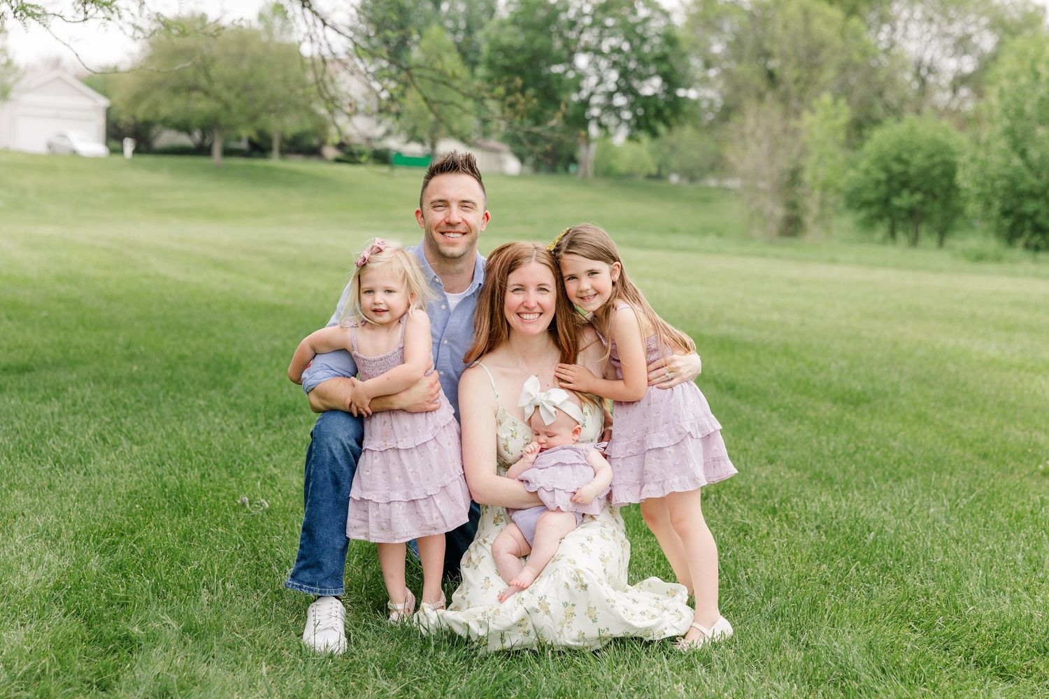 A smiling family of five poses together on a green lawn, with parents and three young daughters.