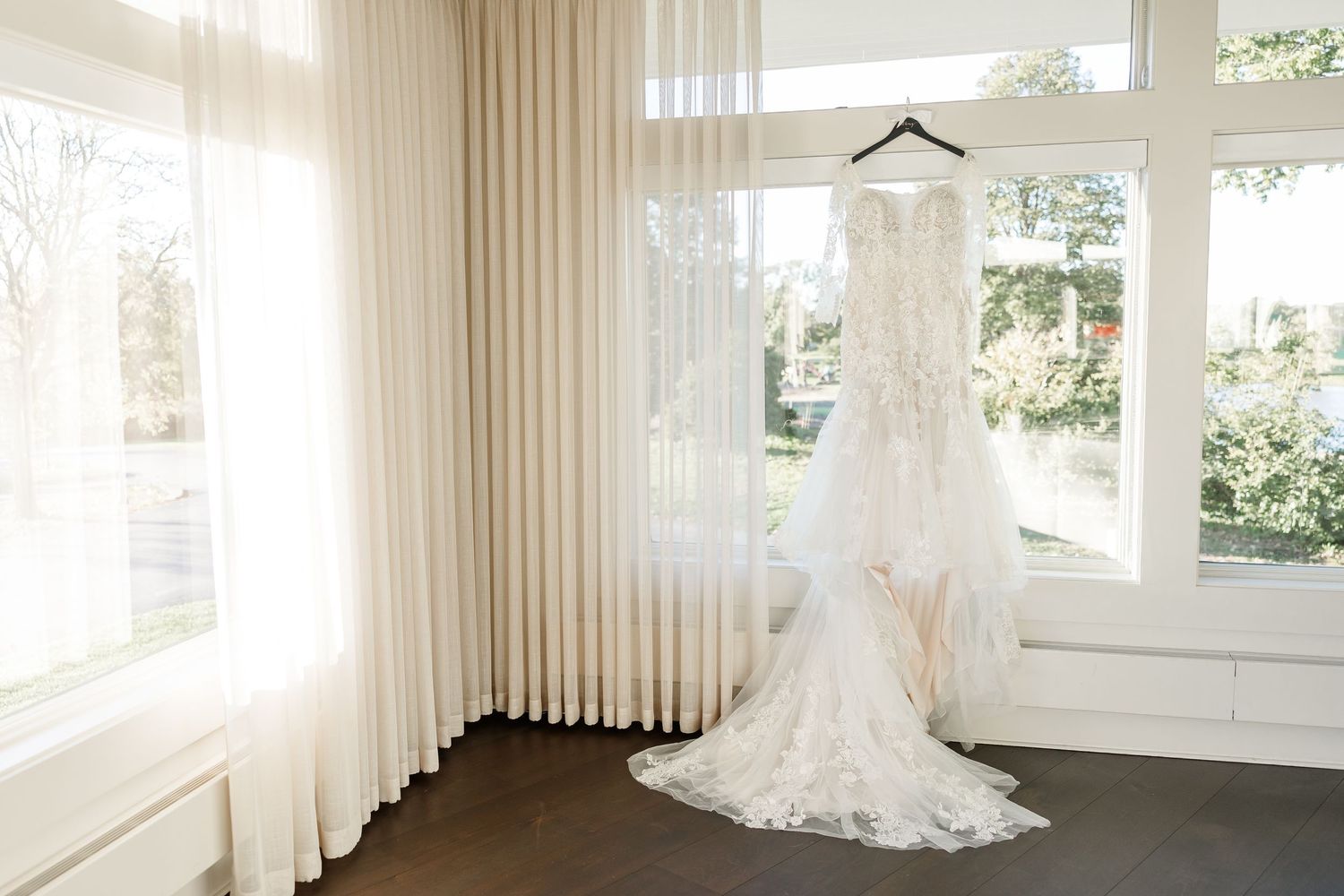 White lace wedding gown with a long flowing train hanging by a bright window with sheer cream curtains in an elegant bridal suite.