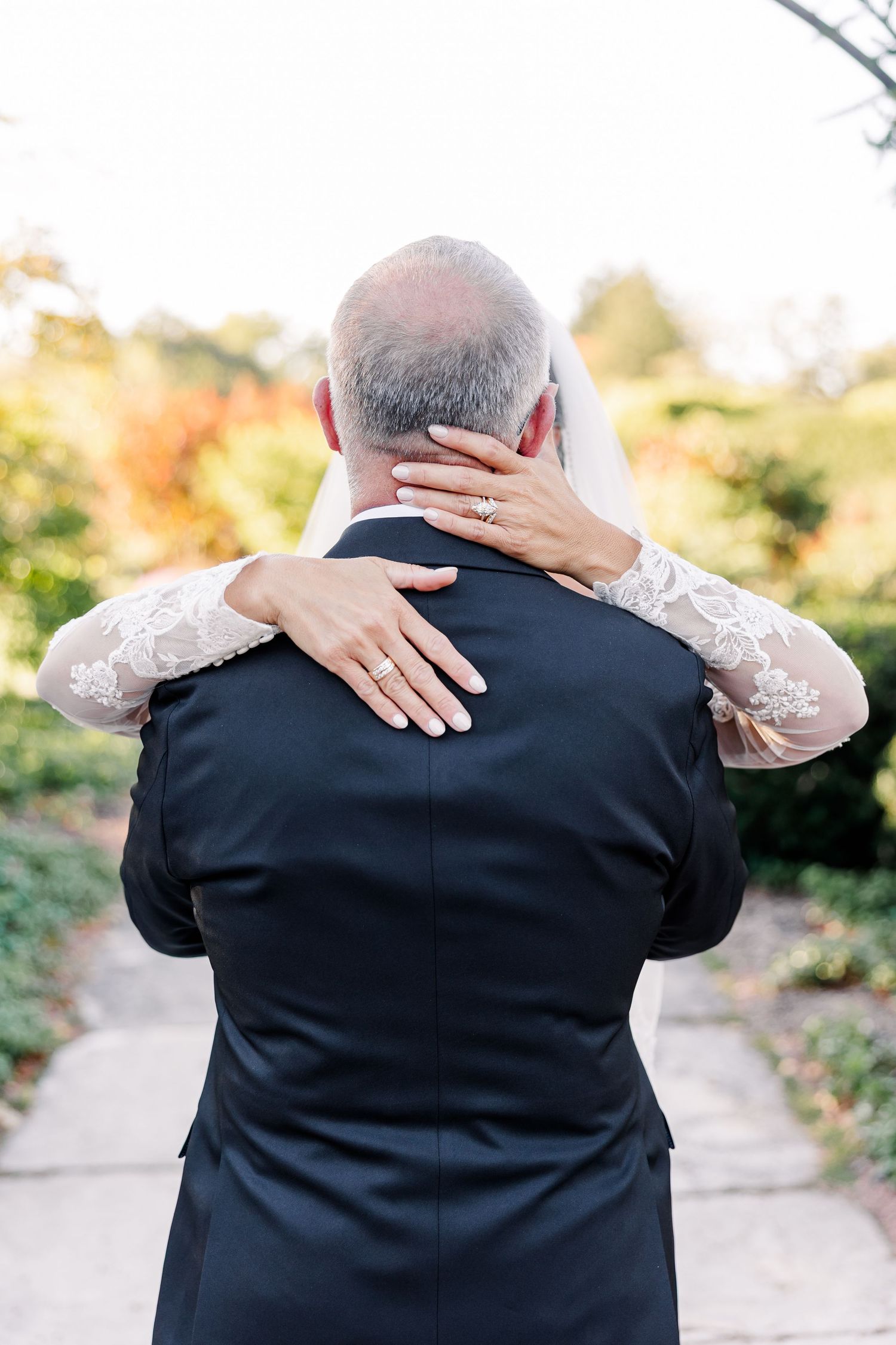 Bride in a lace-sleeved gown and veil embraces a gray-haired groom in a dark suit on a garden path during outdoor wedding portraits.