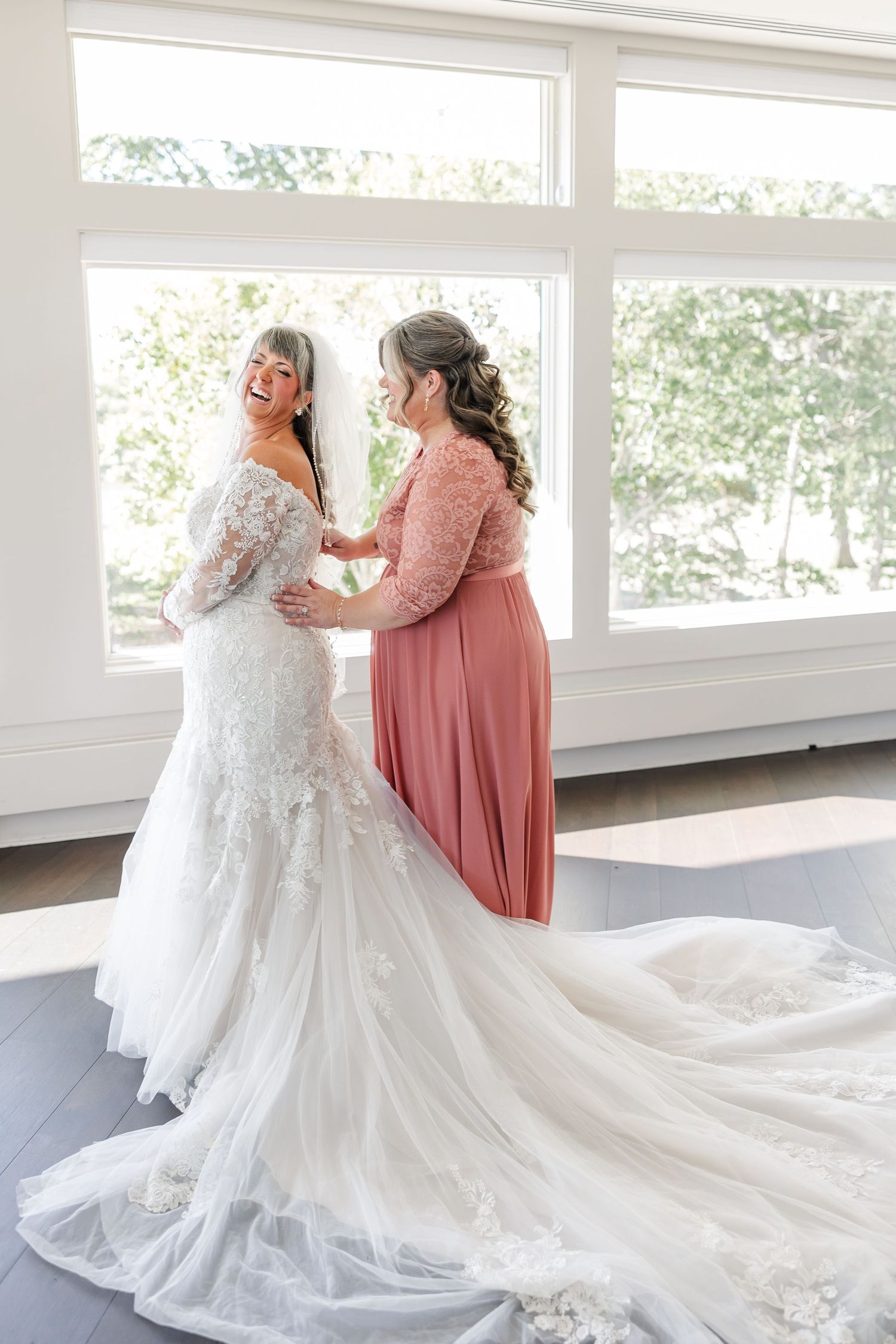 Laughing bride in a lace mermaid gown with a flowing tulle train is helped by a bridesmaid in a coral dress before the ceremony.