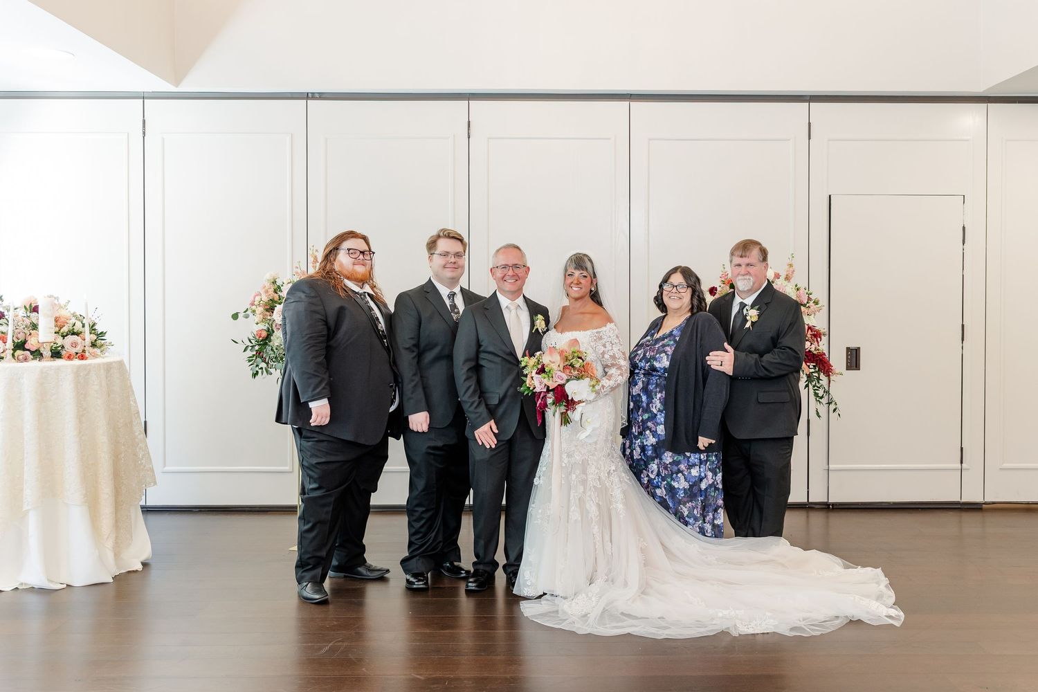A bride in a white gown holding a bouquet poses with five family members in formal attire inside a bright wedding venue.