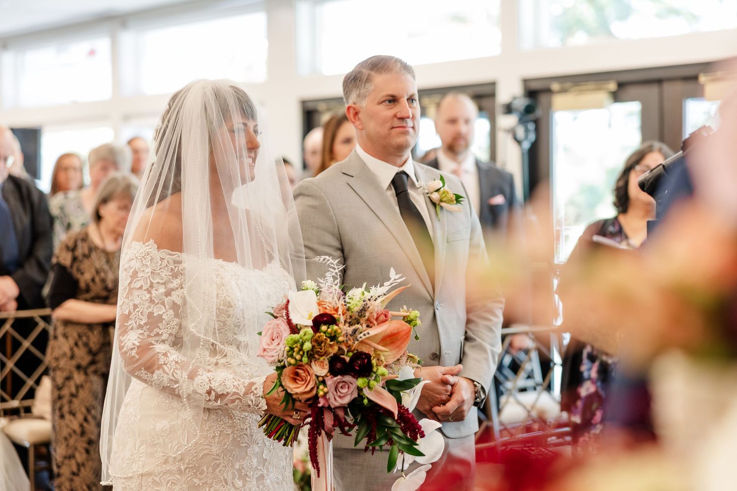 Bride in a lace gown and veil holds a colorful bouquet while standing beside a groom in a gray suit during a wedding ceremony with guests.