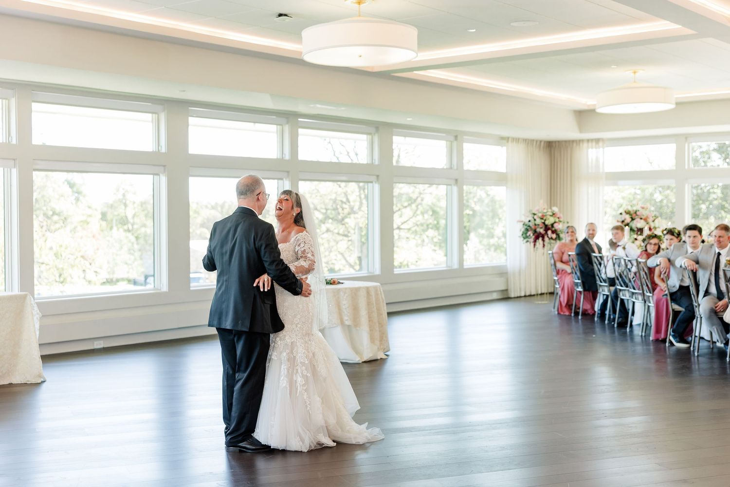 Bride and groom share a first dance on a hardwood floor in a bright reception hall with large windows as guests watch from chairs.