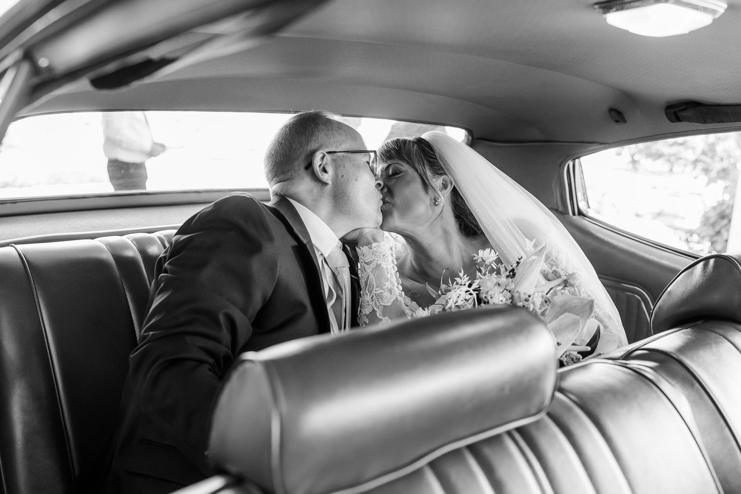 Bride in a veil and groom in a suit share a kiss in the back seat of a vintage car in a black and white wedding photo.
