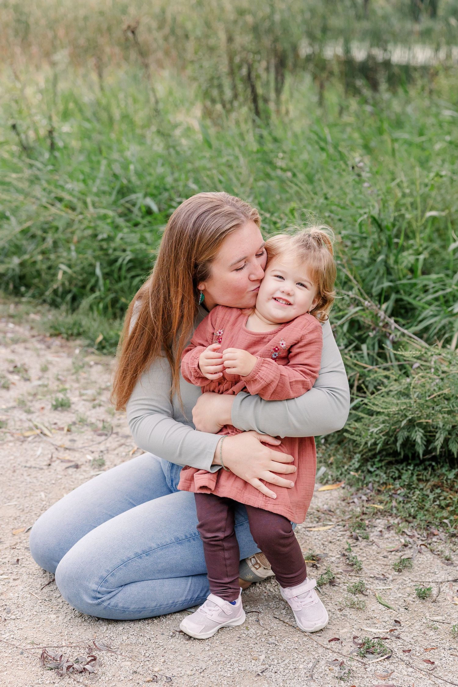 Mother kneeling on sandy ground kissing her smiling toddler daughter on the cheek near tall green grass outdoors.
