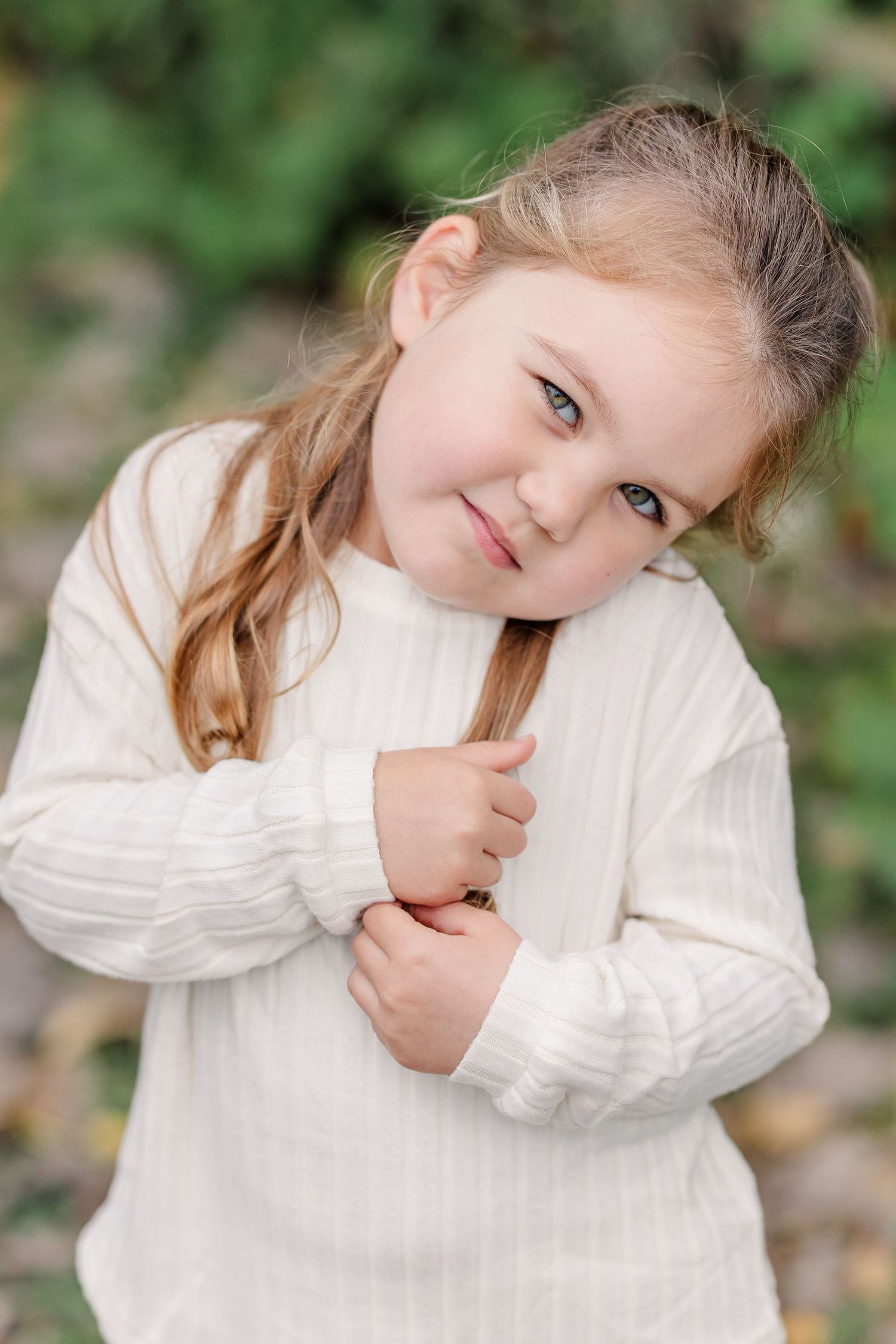 A cute little girl in a cream ribbed sweater tilts her head and smiles softly in a green outdoor setting.