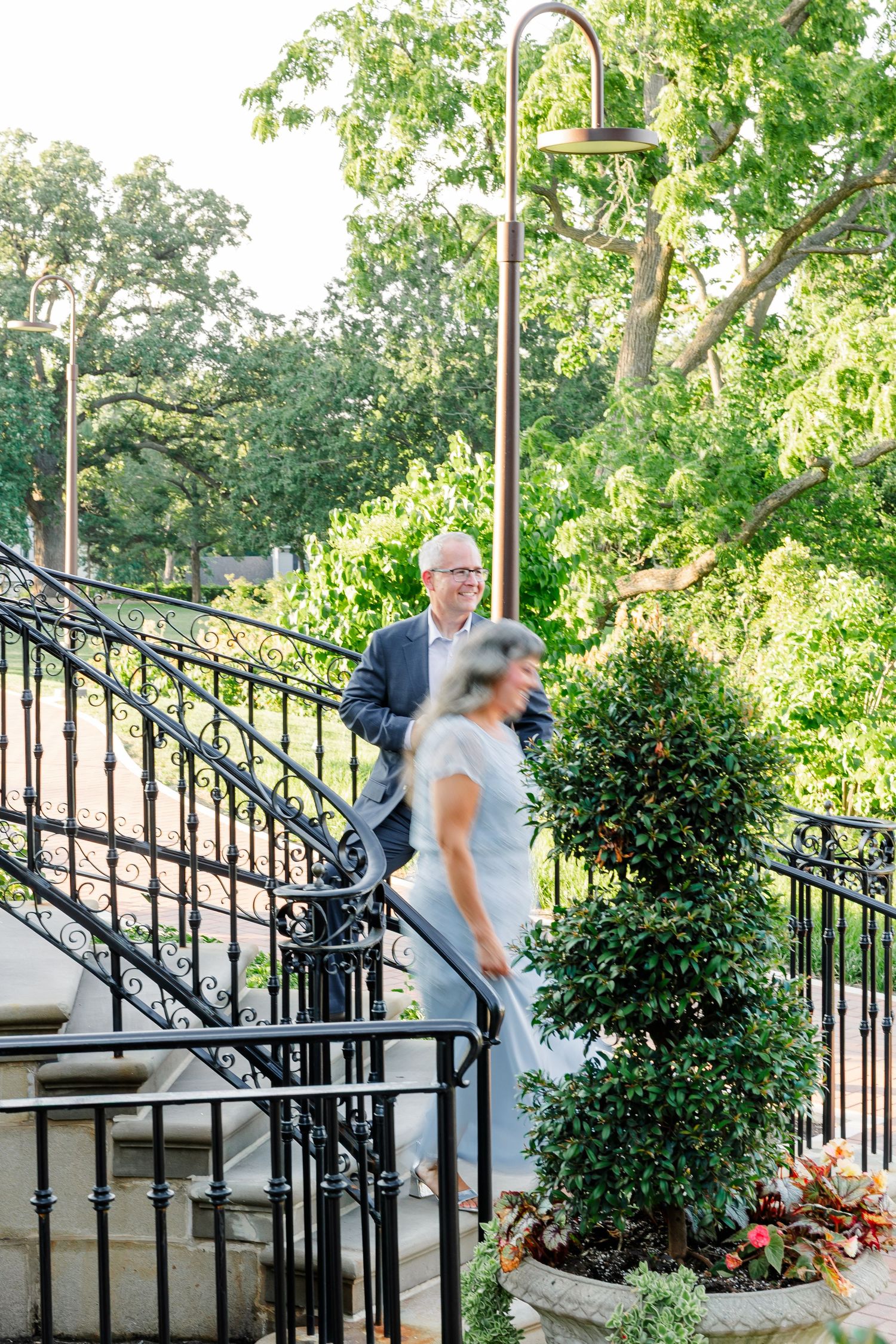 Bride and groom descend elegant iron staircase surrounded by lush greenery at an outdoor wedding venue.