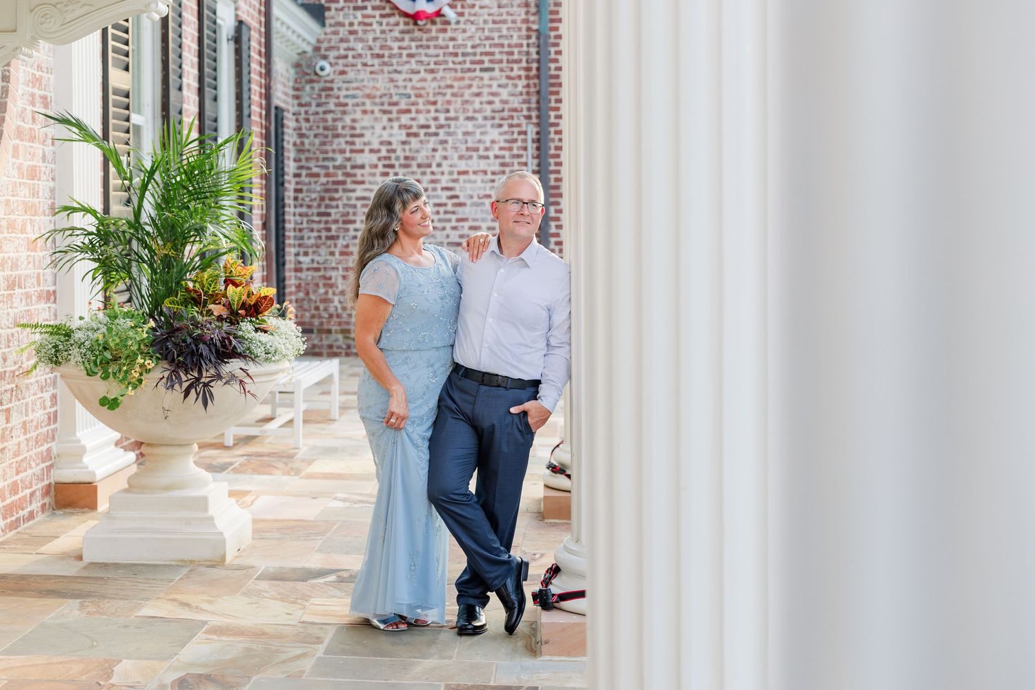 Couple in formal attire walking together on an elegant brick porch with white columns and tropical floral arrangements.