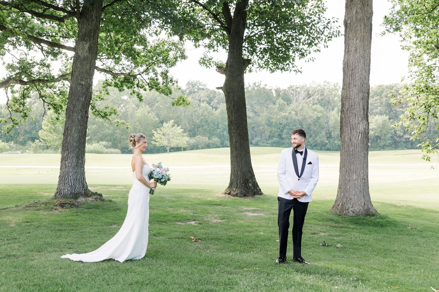 Bride and groom share a first look moment outdoors beneath large trees on a lush green lawn.