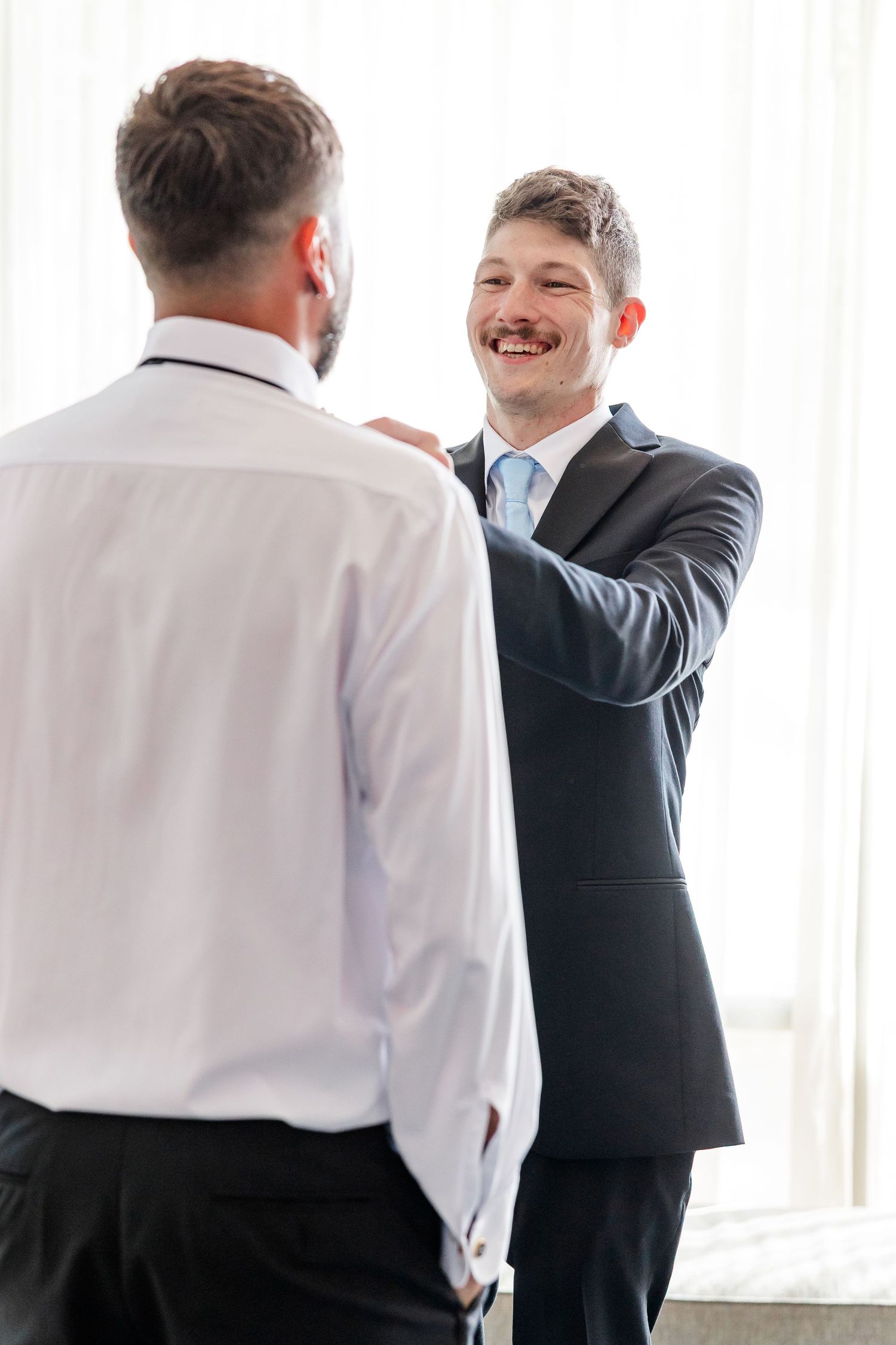 Smiling groomsman in dark suit helps groom adjust tie before wedding ceremony.