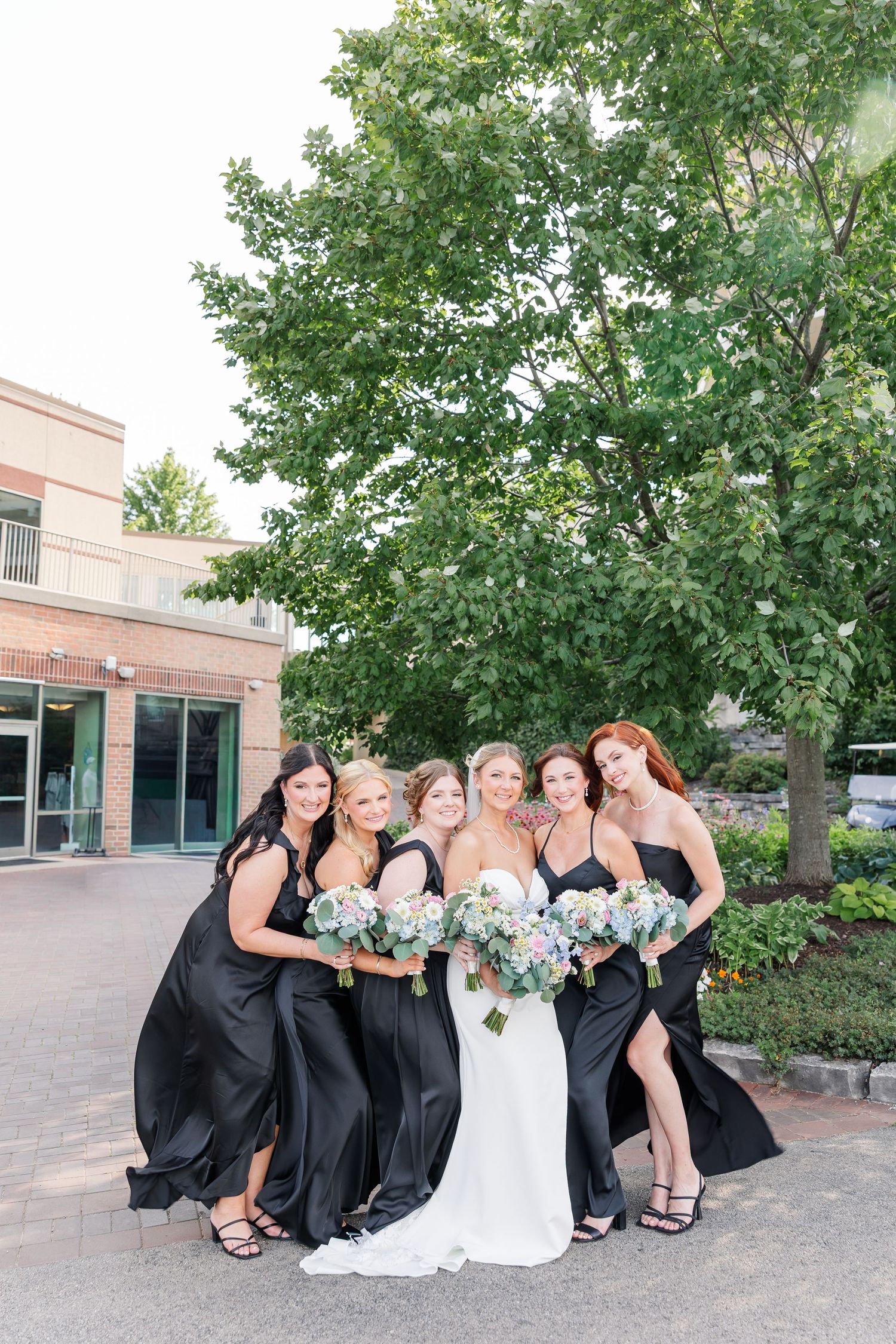 Bride in white gown poses with five bridesmaids in black dresses holding floral bouquets outdoors.
