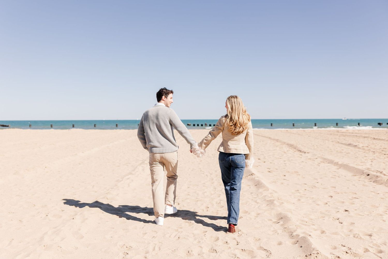 A couple holds hands walking on a sandy beach with calm blue ocean waters and clear sky in the background.