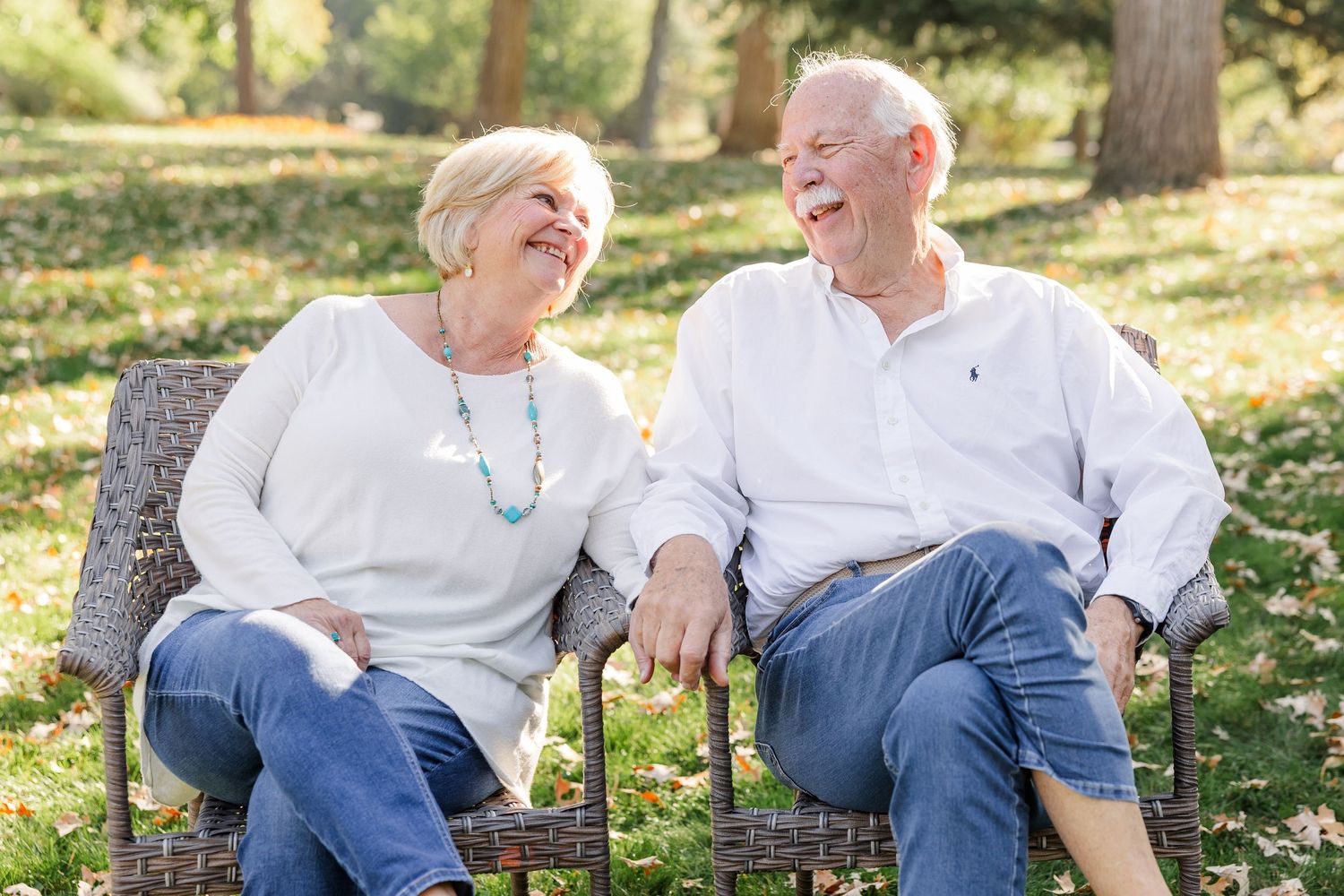 Elderly couple laughing together on a wicker bench in a sunny park, wearing casual jeans and light tops.