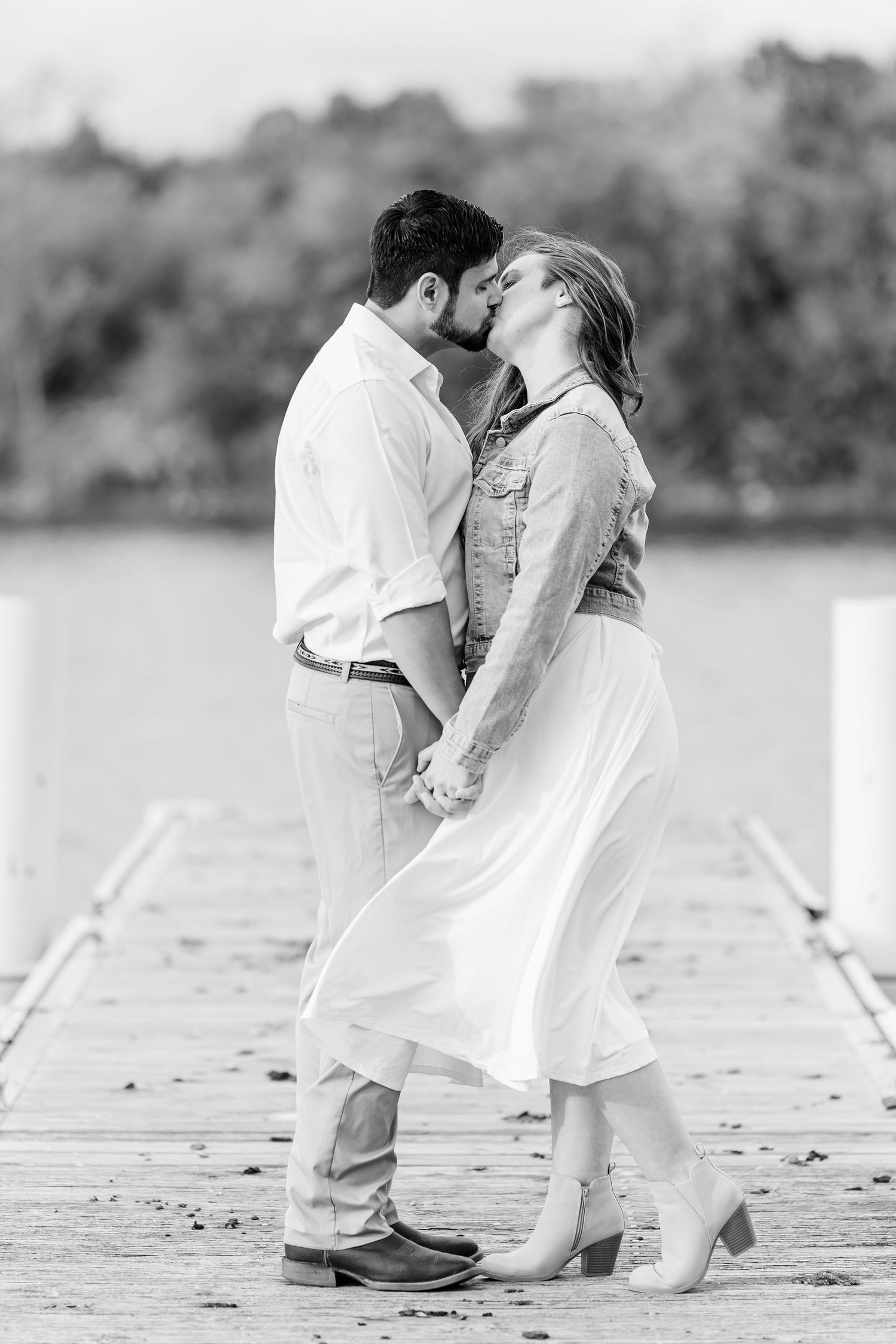 Black and white photo of a couple in a denim jacket and white dress kissing on a wooden dock beside a calm lake.