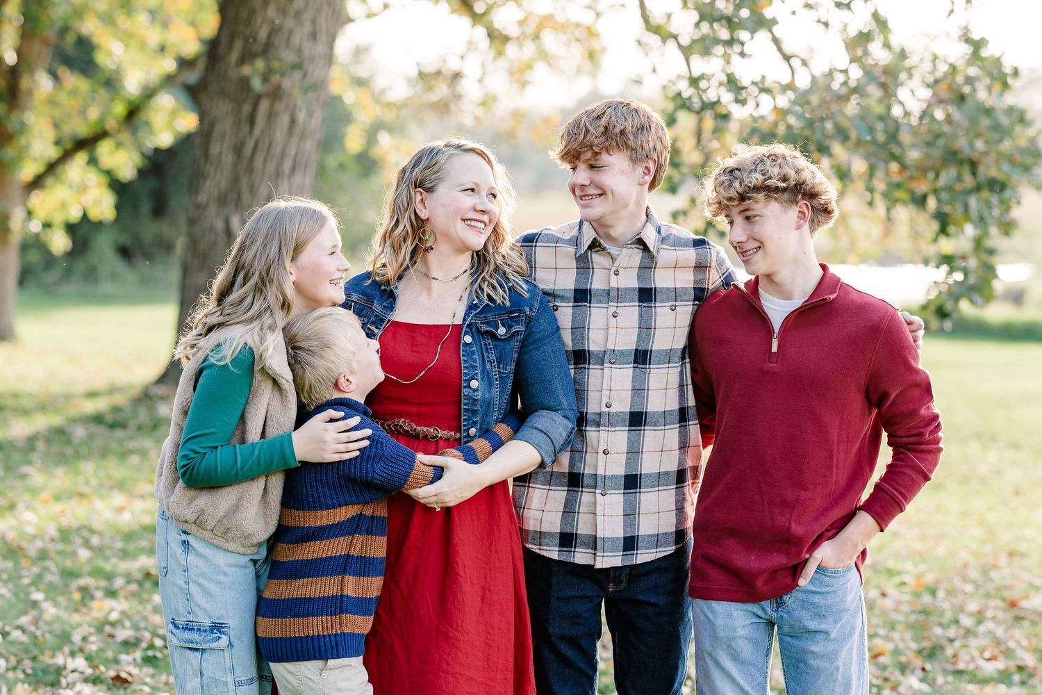 Smiling family of five in coordinated fall outfits stands together outdoors under trees during a sunny autumn family portrait session.