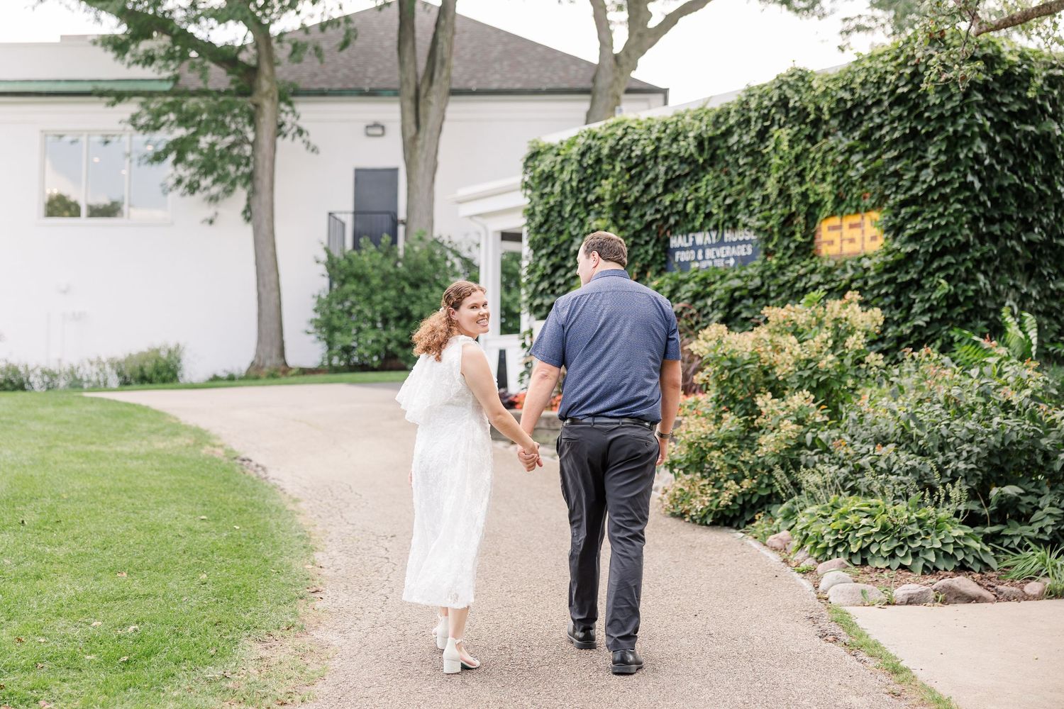 Couple holding hands walking along a garden path outside a white building with ivy walls.